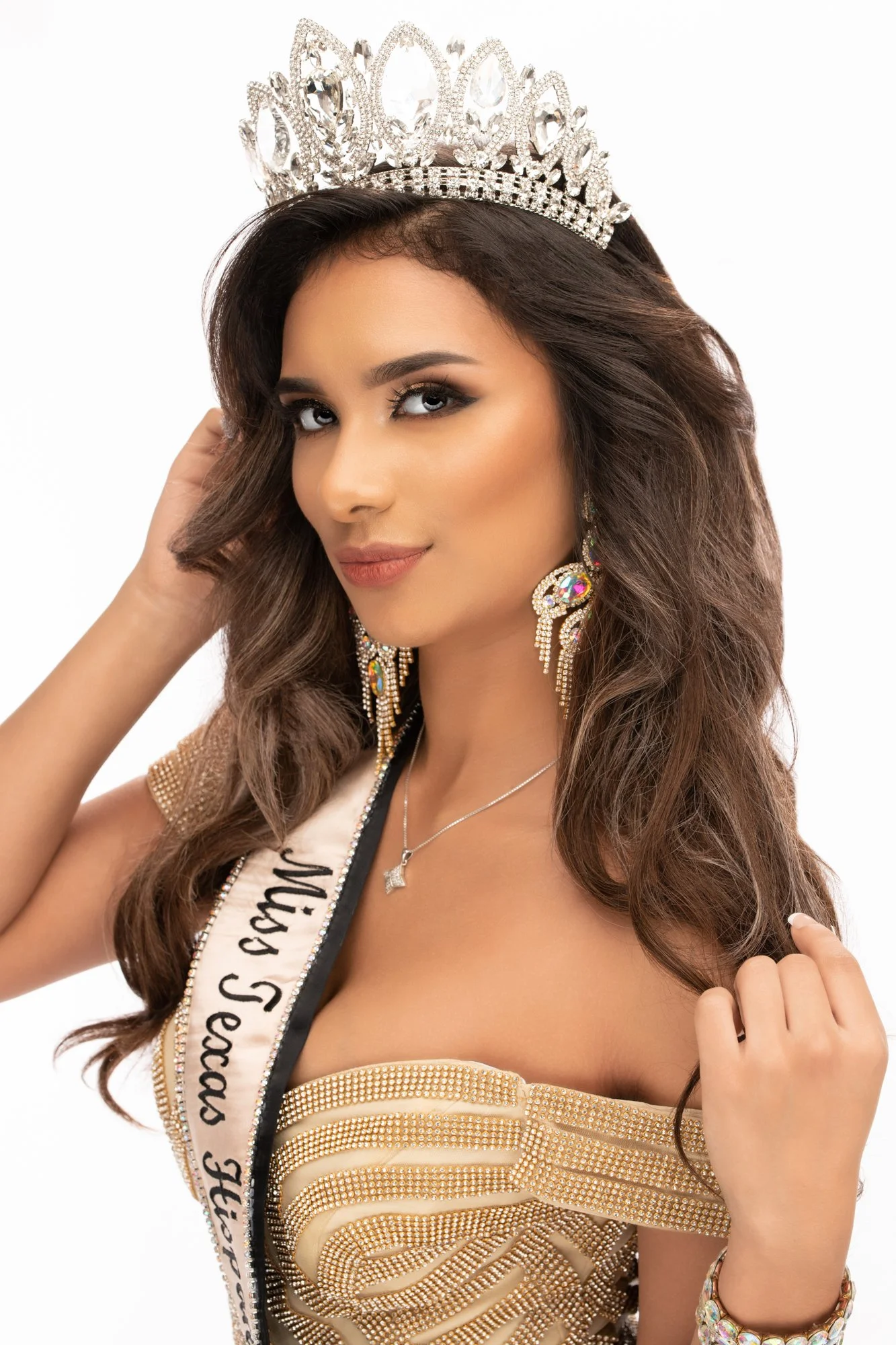 A studio portrait of a pageant winner wearing a crystal crown and an ornate gold gown with a sash, styled with glamorous makeup and statement earrings, posing confidently against a white backdrop.