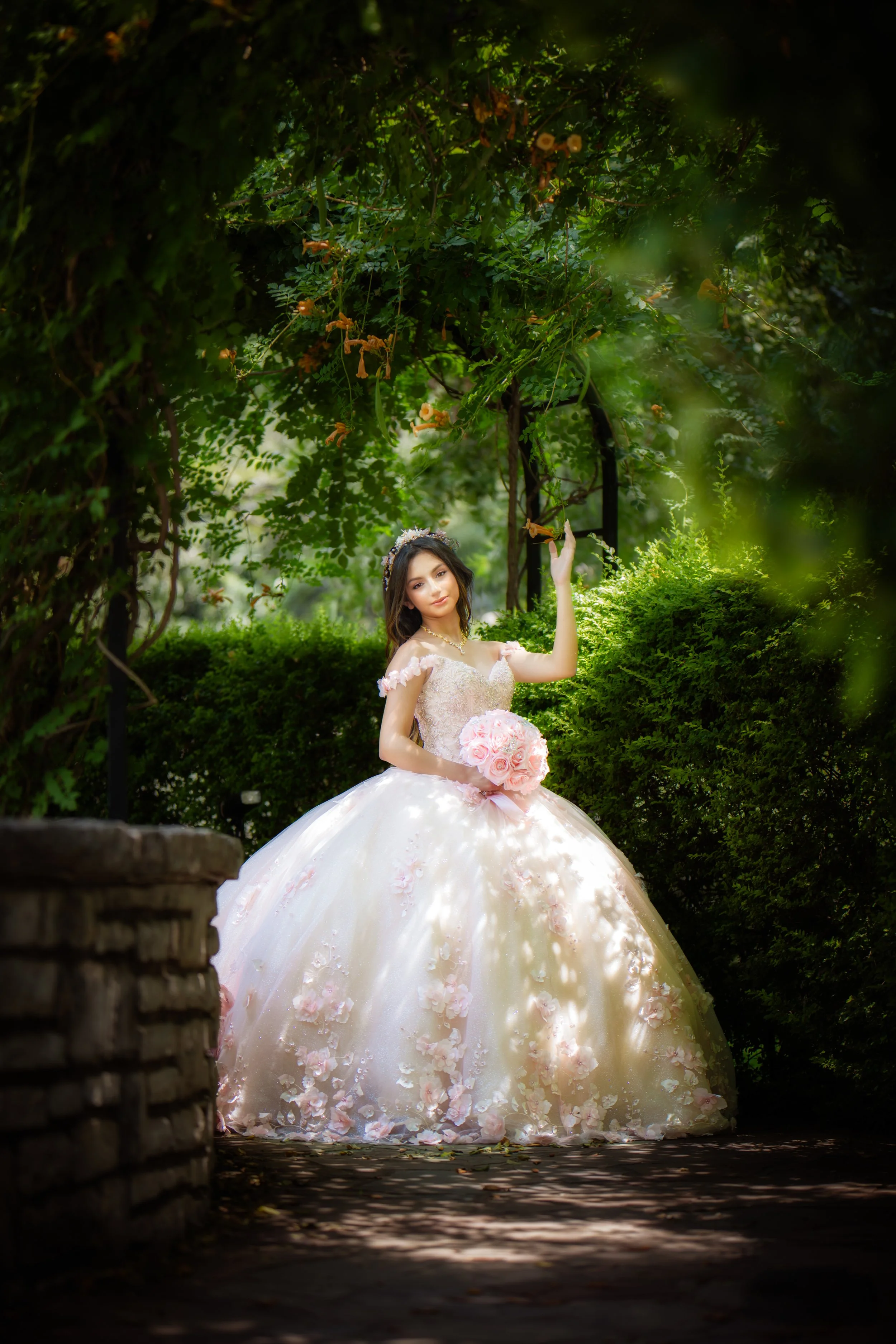 The quinceañera poses gracefully beneath a garden archway, wearing a pastel pink floral gown and holding a rose bouquet, captured in dappled sunlight that highlights her beauty and feminine elegance.