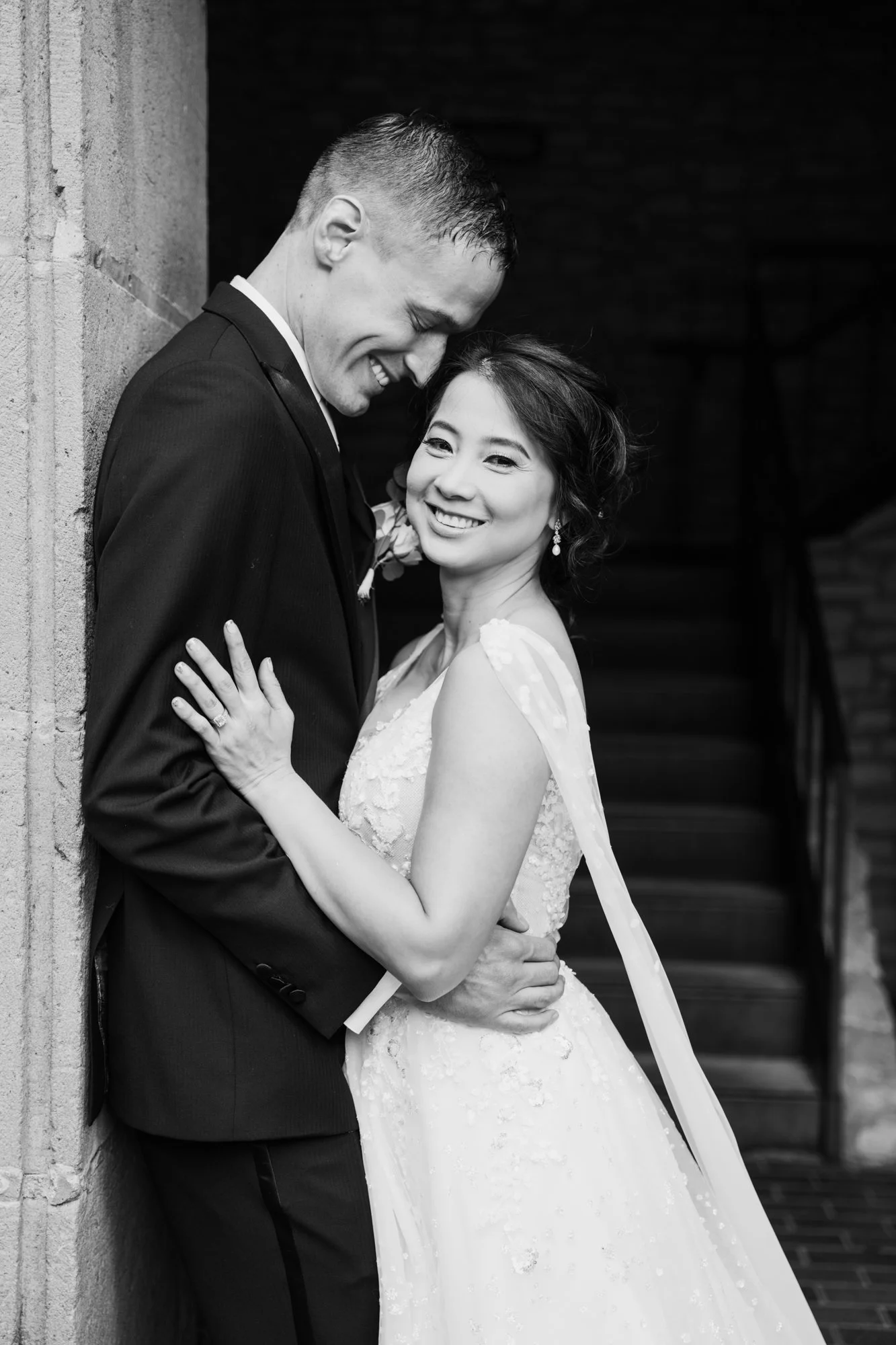 A black-and-white portrait of a newlywed couple standing closely together beneath a stone archway. The groom gently leans his head toward the bride while she smiles directly at the camera, her veil softly draped behind her, capturing an intimate and 