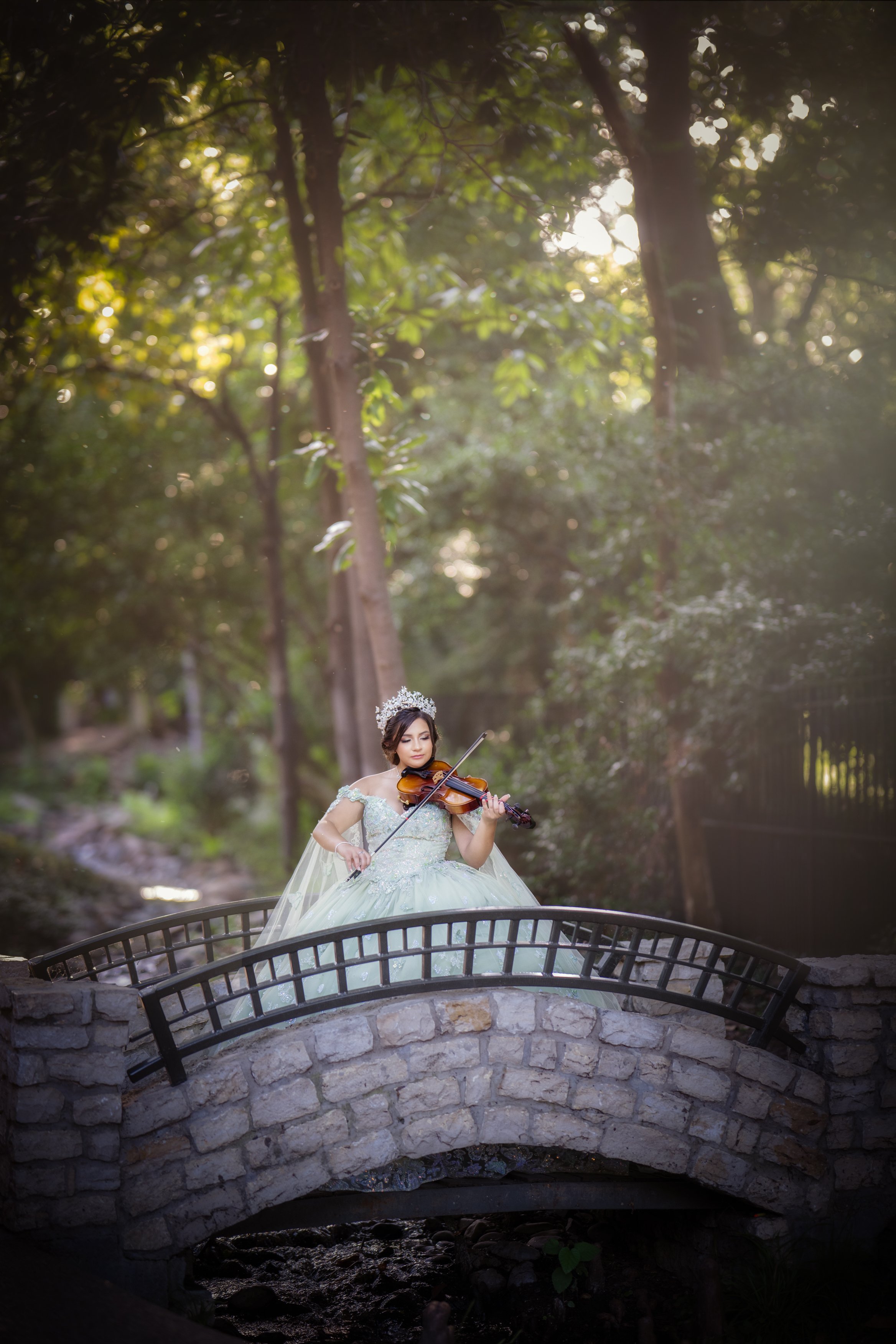 A quinceañera plays the violin while standing on a stone bridge in a lush park, her mint green gown flowing gracefully as soft afternoon light filters through the trees, creating a serene and artistic moment.