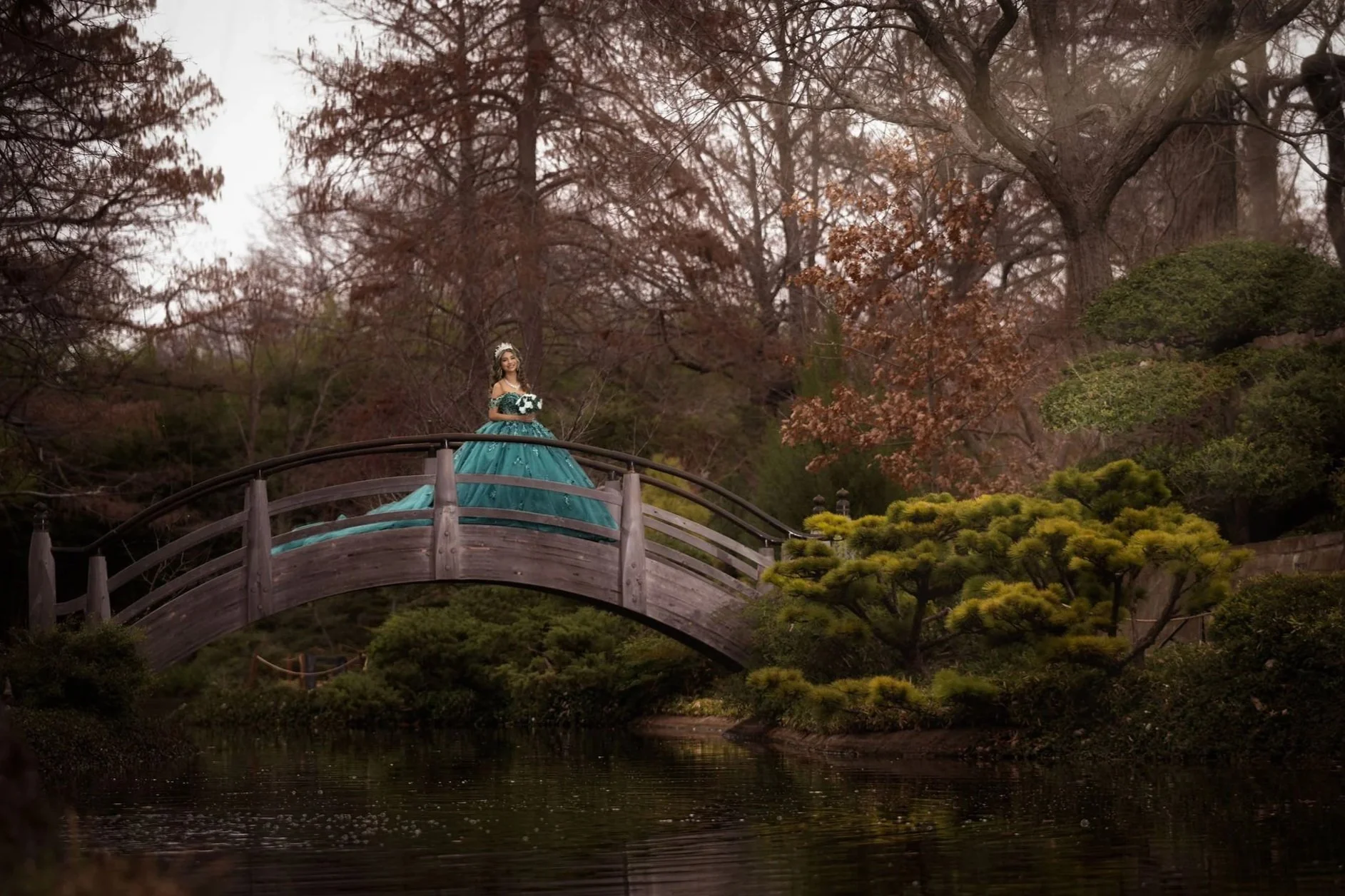 A woman in a teal ball gown and tiara stands on a wooden arched bridge over a pond in a garden with trees and shrubs.