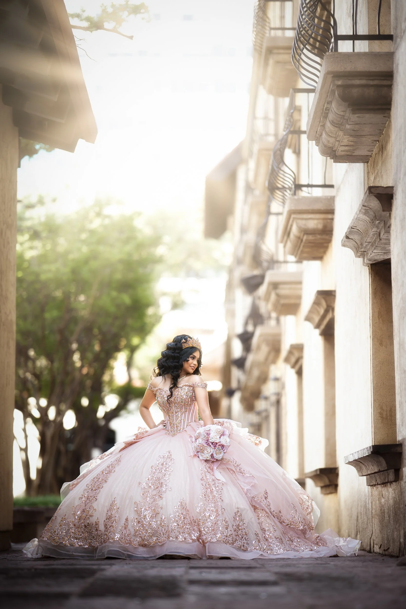 A wide architectural portrait captures the quinceañera centered beneath a grand wooden gate, framed by natural greenery. Her blush pink quinceañera dress flows outward, creating a dramatic and regal composition.