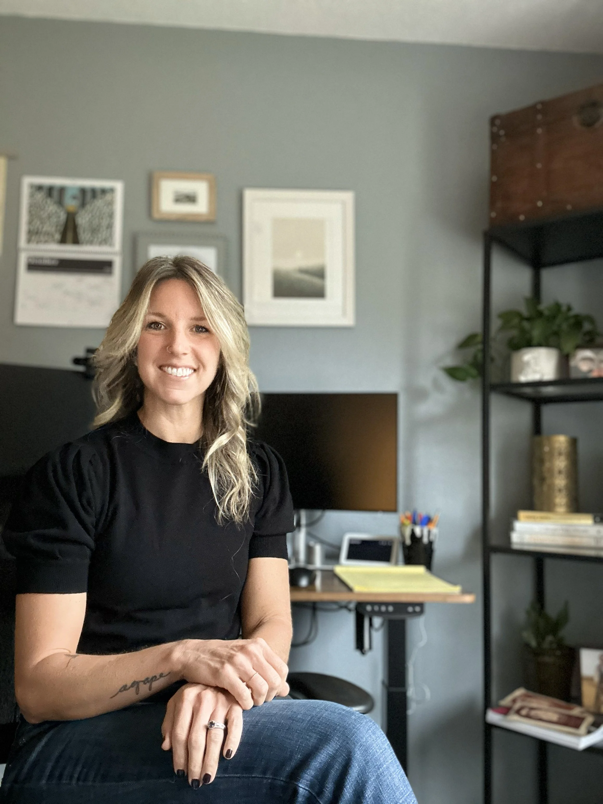 A woman sitting in a home office, smiling at the camera, with a light blue wall, framed pictures, a computer monitor, a desk with office supplies, and a black bookshelf with plants and books in the background.