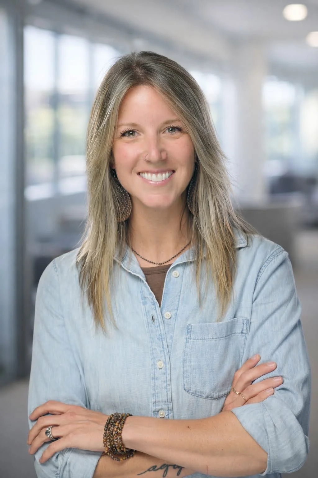 A woman with long, blonde hair and a nose piercing smiling, standing cross-armed in a modern office setting with large windows and blurred background.