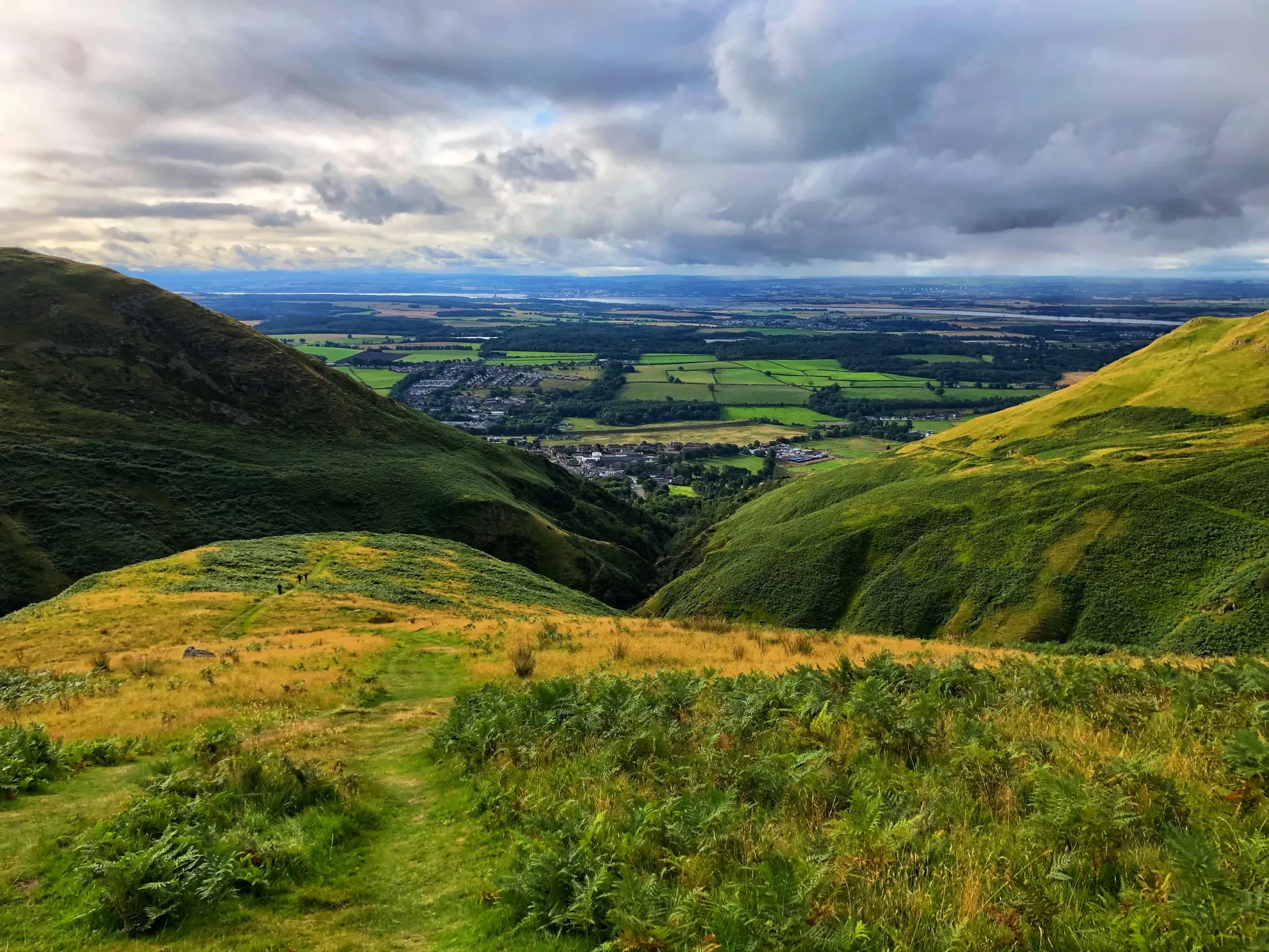 A scenic view of green rolling hills and a valley with fields, trees, and buildings, under a cloudy sky.