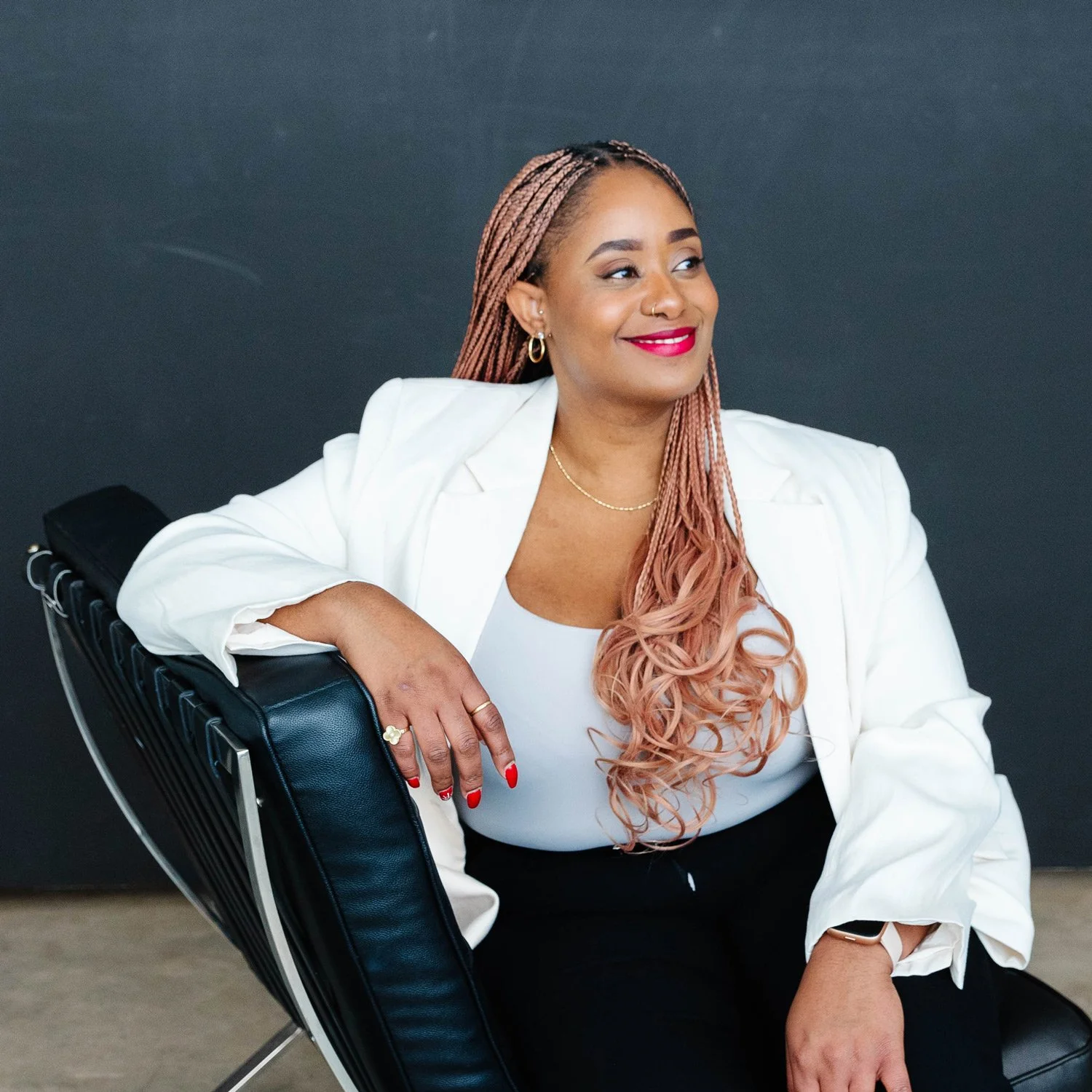 Smiling woman with long pink braids wearing a white blazer seated in a black leather chair