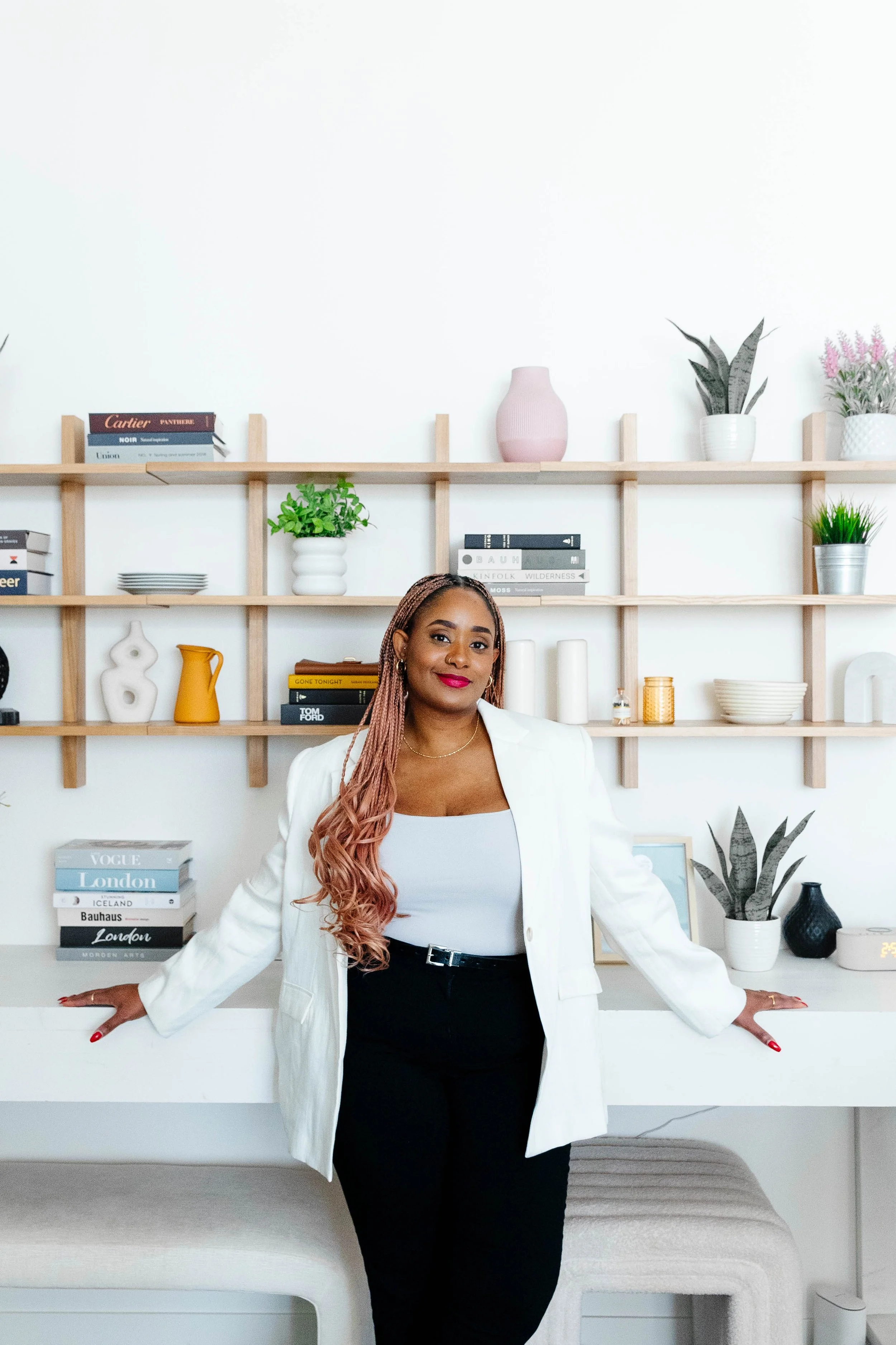 Smiling woman in a white blazer and black pants standing in front of minimalist shelves with books, plants, and decorative objects