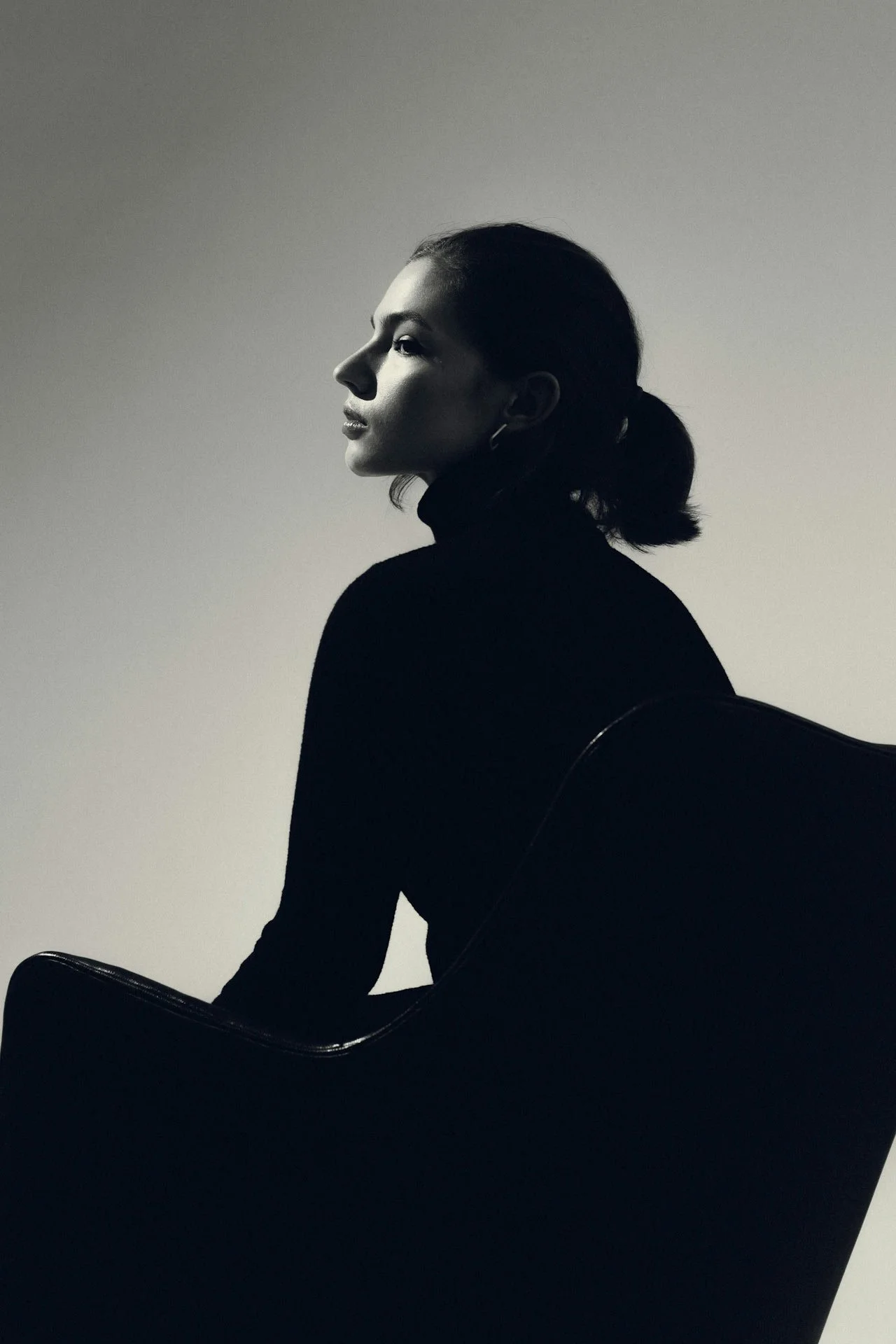 Black-and-white side-profile portrait of a woman with a ponytail seated in a dark chair