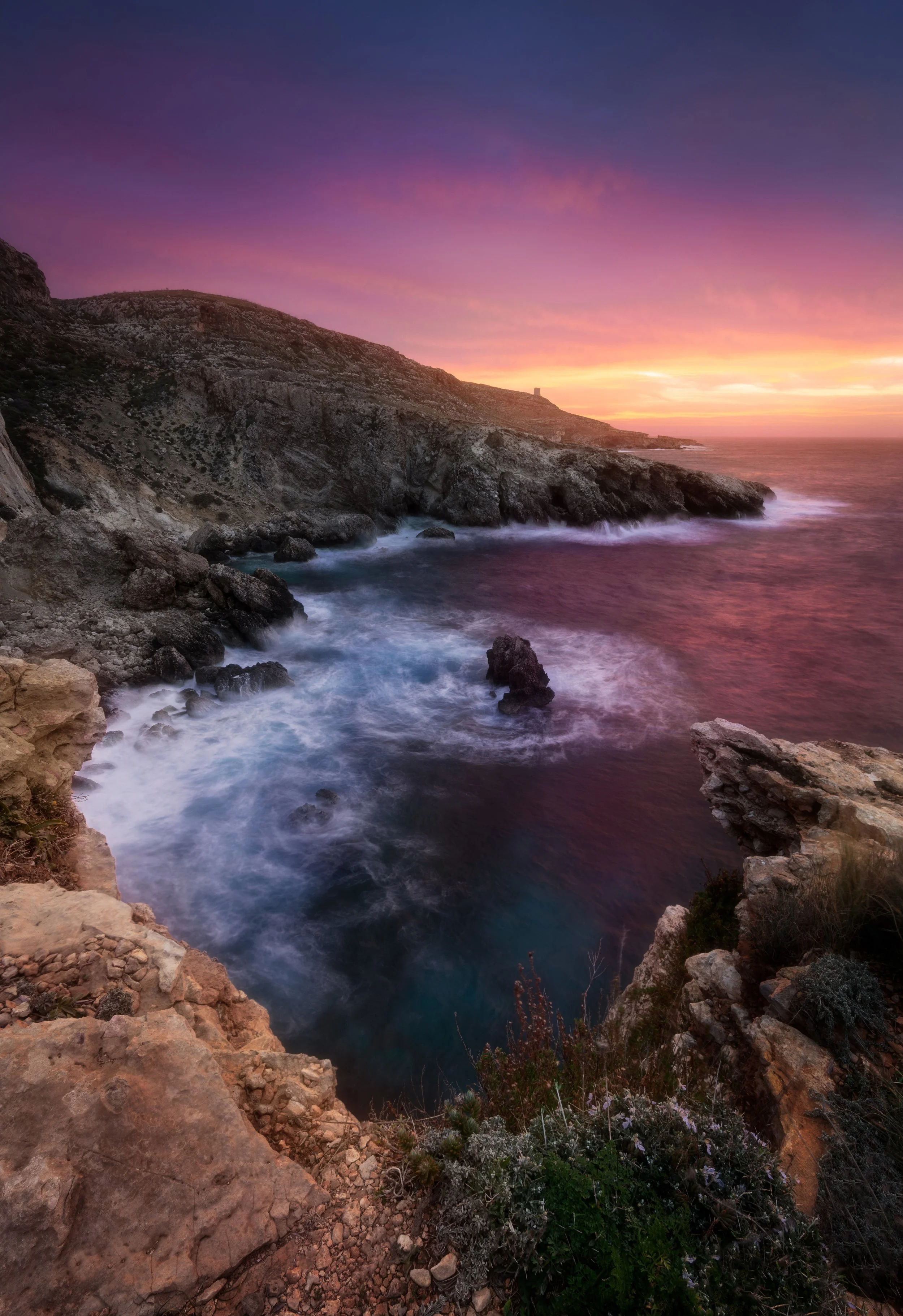 A coastal landscape during sunset with rocky cliffs, ocean waves, and a colorful sky with shades of pink, purple, and orange.