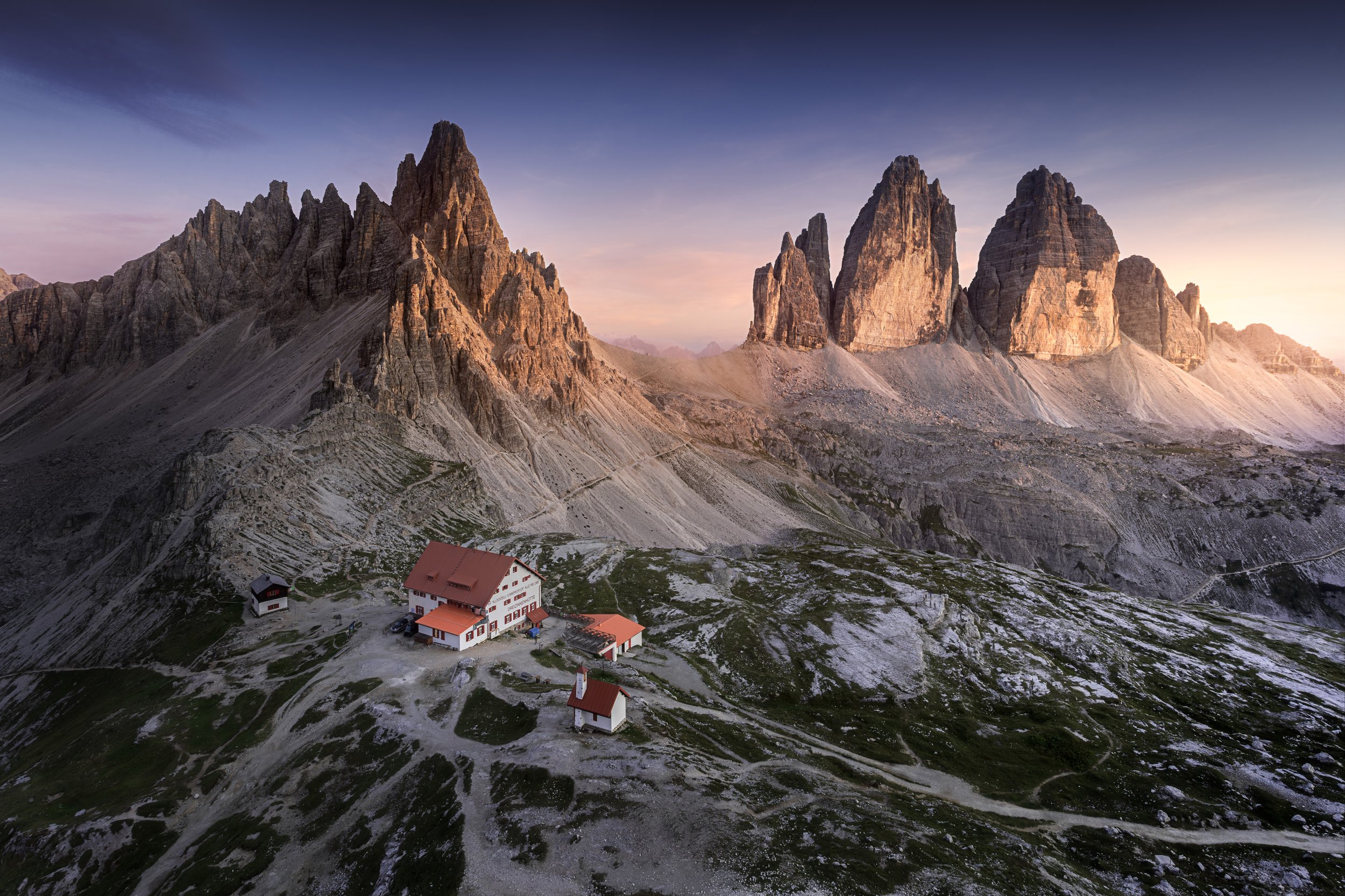 A mountain landscape featuring jagged peaks with a small mountain hut and church with red roofs in the foreground, during sunset or sunrise.