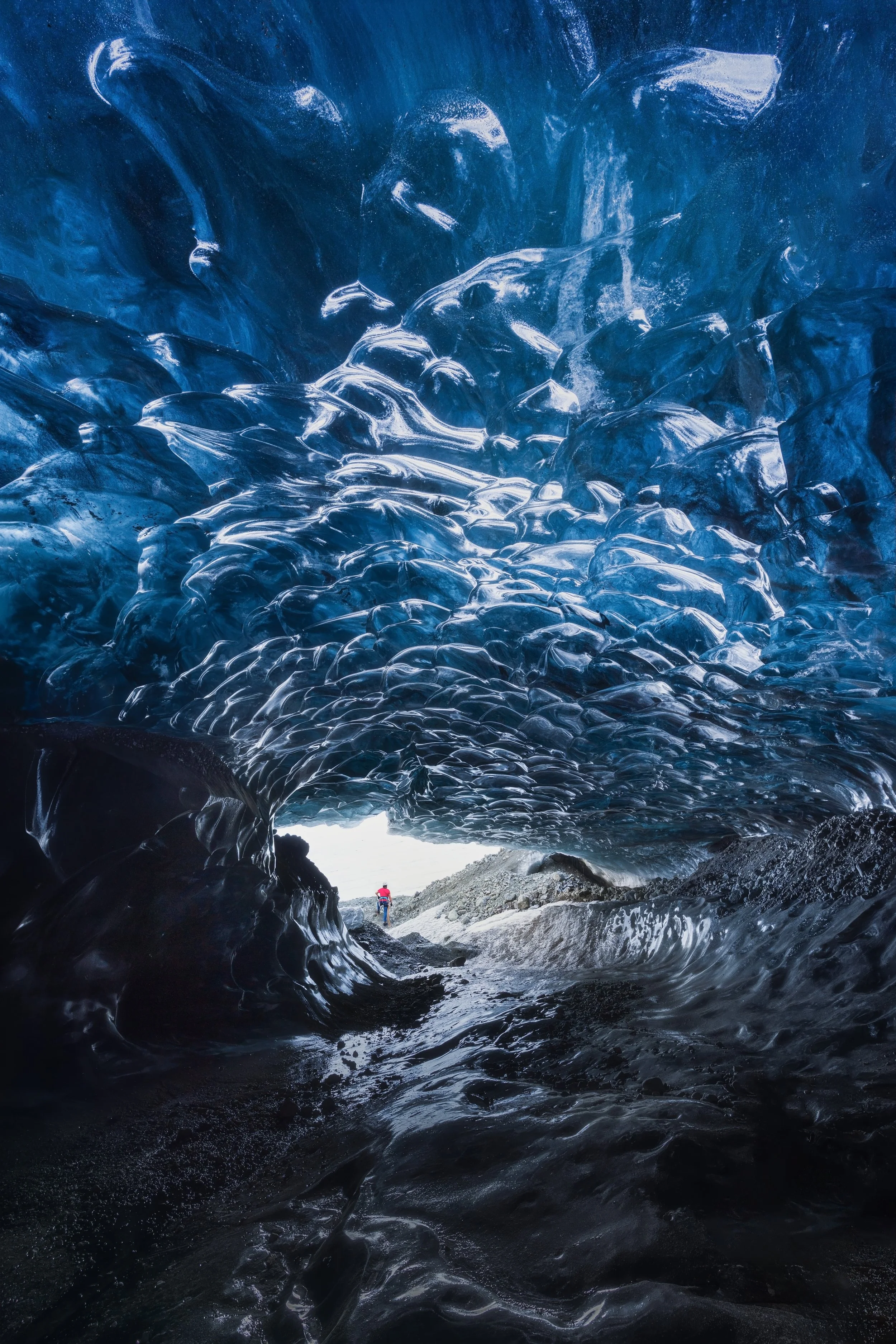 View from inside an ice cave showing intricate blue ice formations and a person standing outside at the cave entrance.