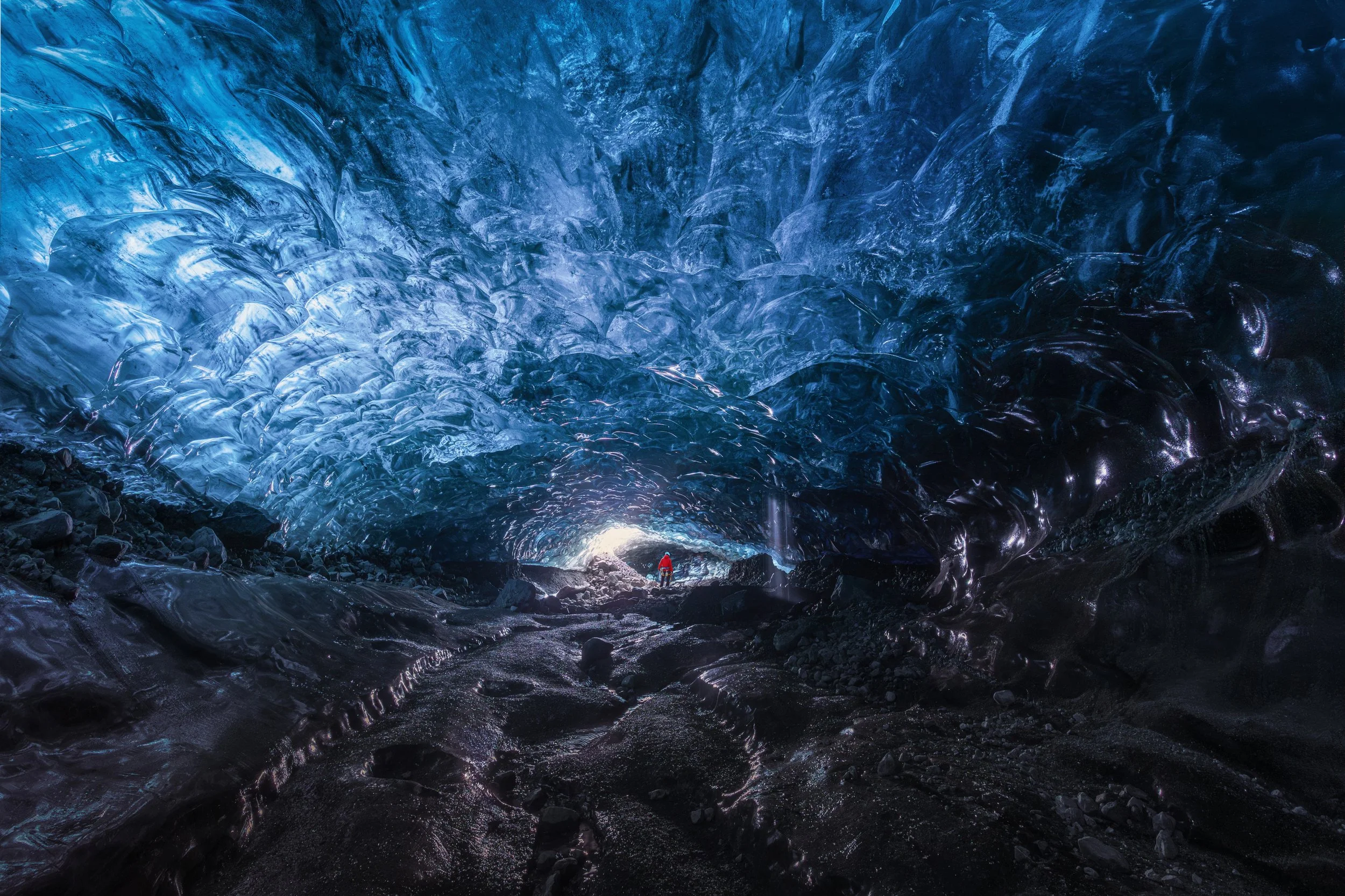 A person in a red jacket standing inside an ice cave with blue and black ice formations on the ceiling and walls, and dark rocky ground.
