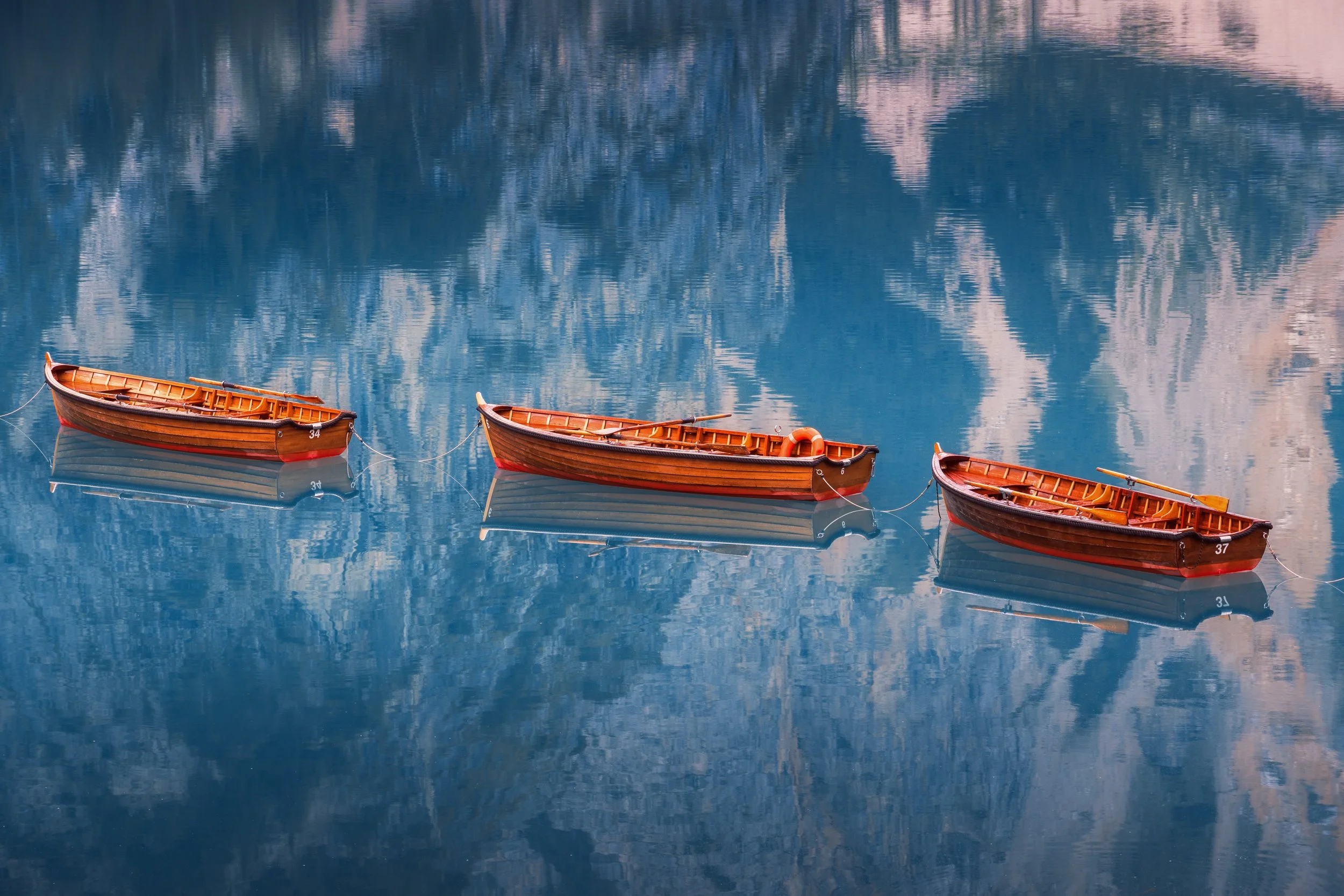 Three small wooden rowboats anchored in calm water, with reflections of mountains and sky visible in the water.