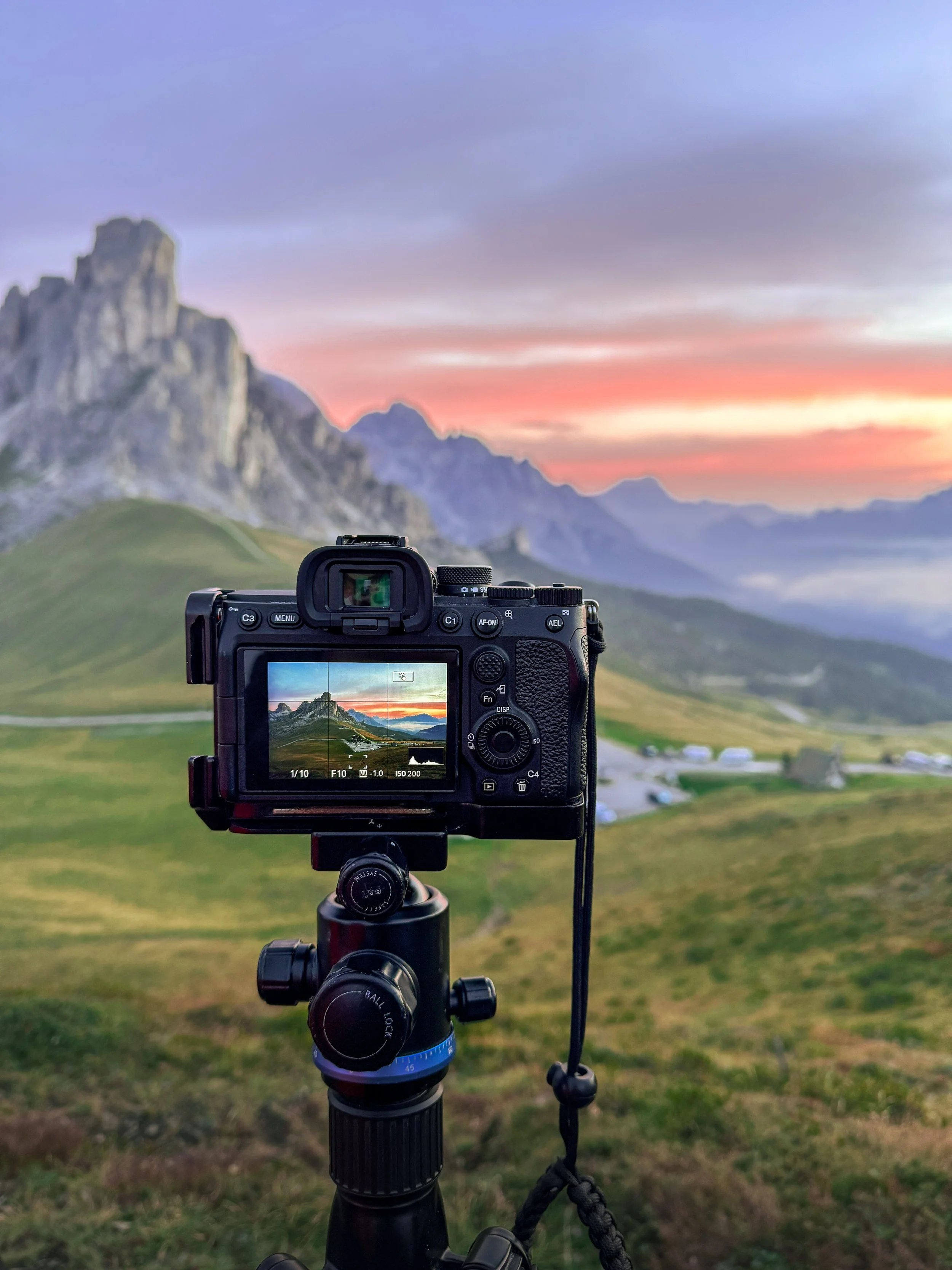 A camera on a tripod captures a landscape scene of rolling green hills, rugged mountains, and a colorful sunset sky.