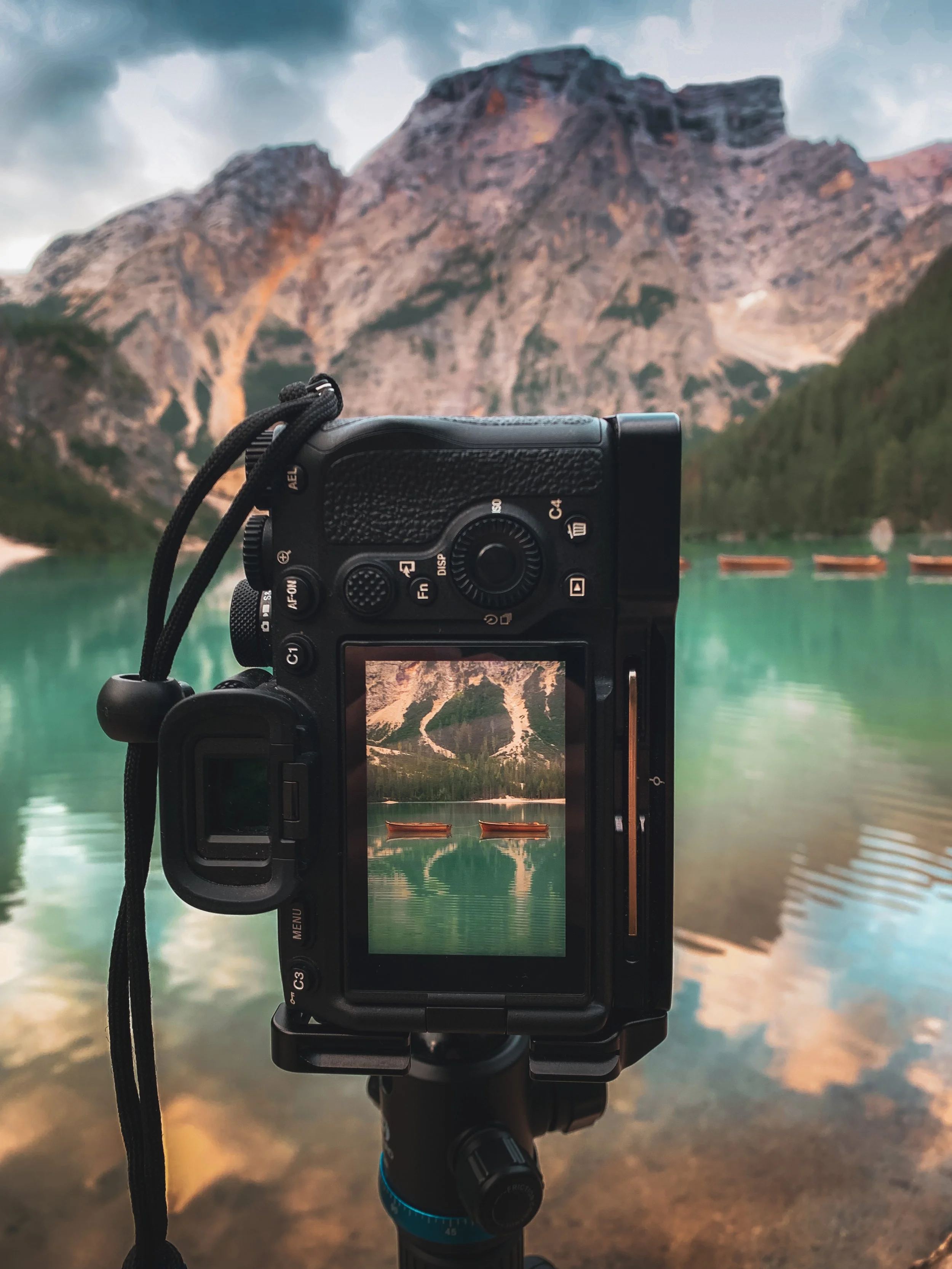 A camera on a tripod capturing a landscape of a mountain lake with green water and boats, mountains, and sky in the background.