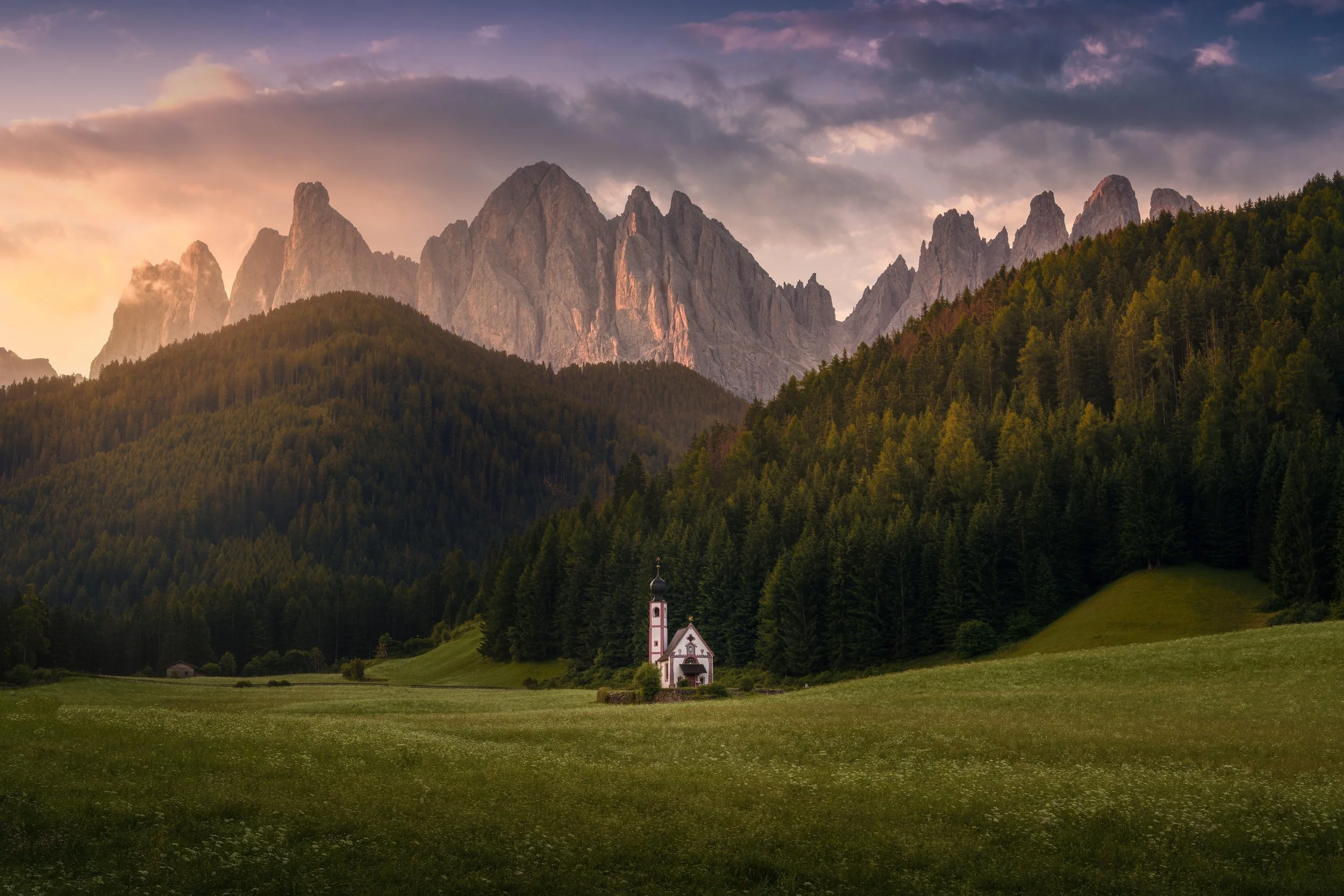 A small white church with a bell tower in a green meadow, surrounded by a forest with mountain peaks in the background during sunset.
