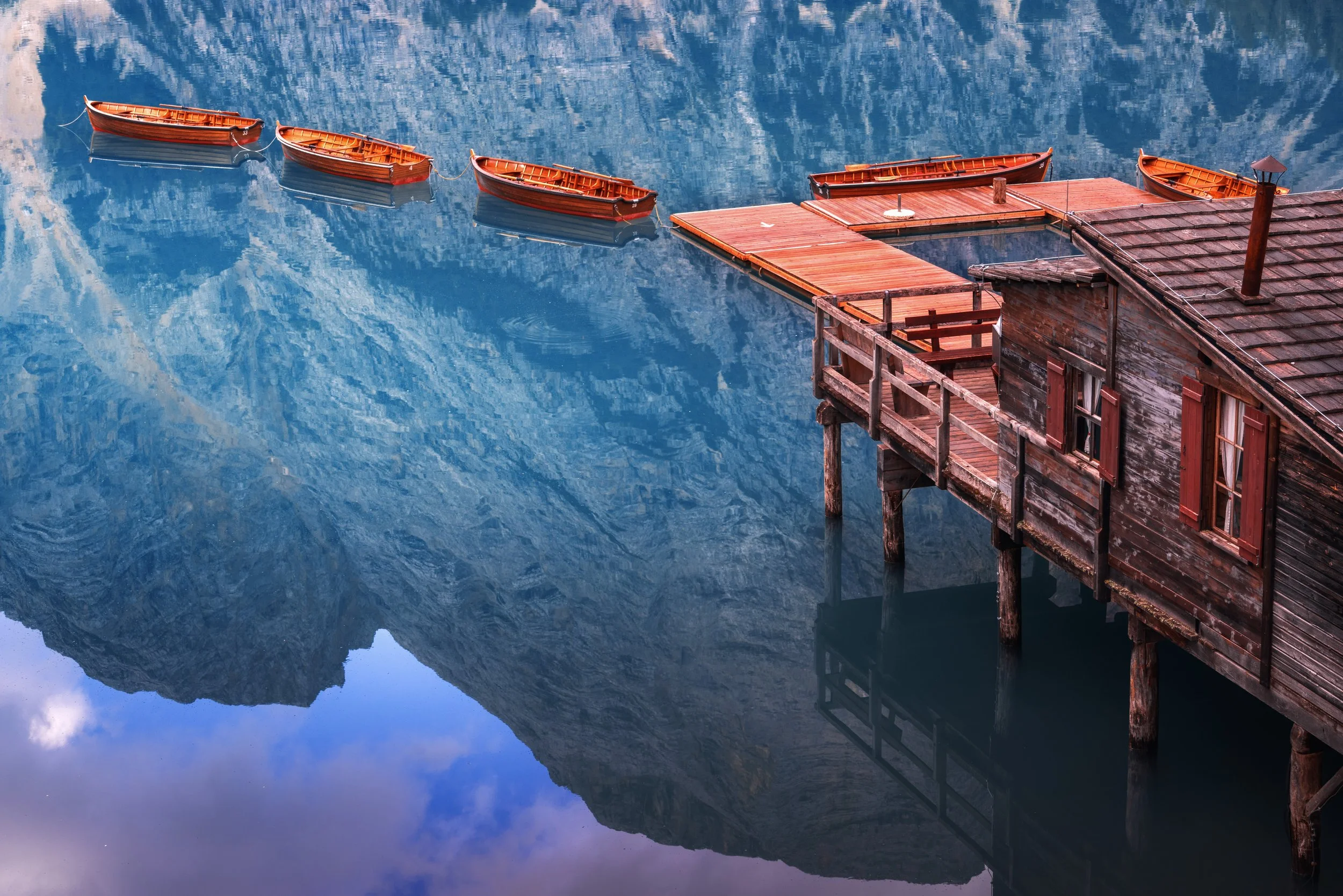 A wooden cabin with a deck extending over the water, and four small wooden boats tied to a floating dock, with a mountain reflected in the calm lake