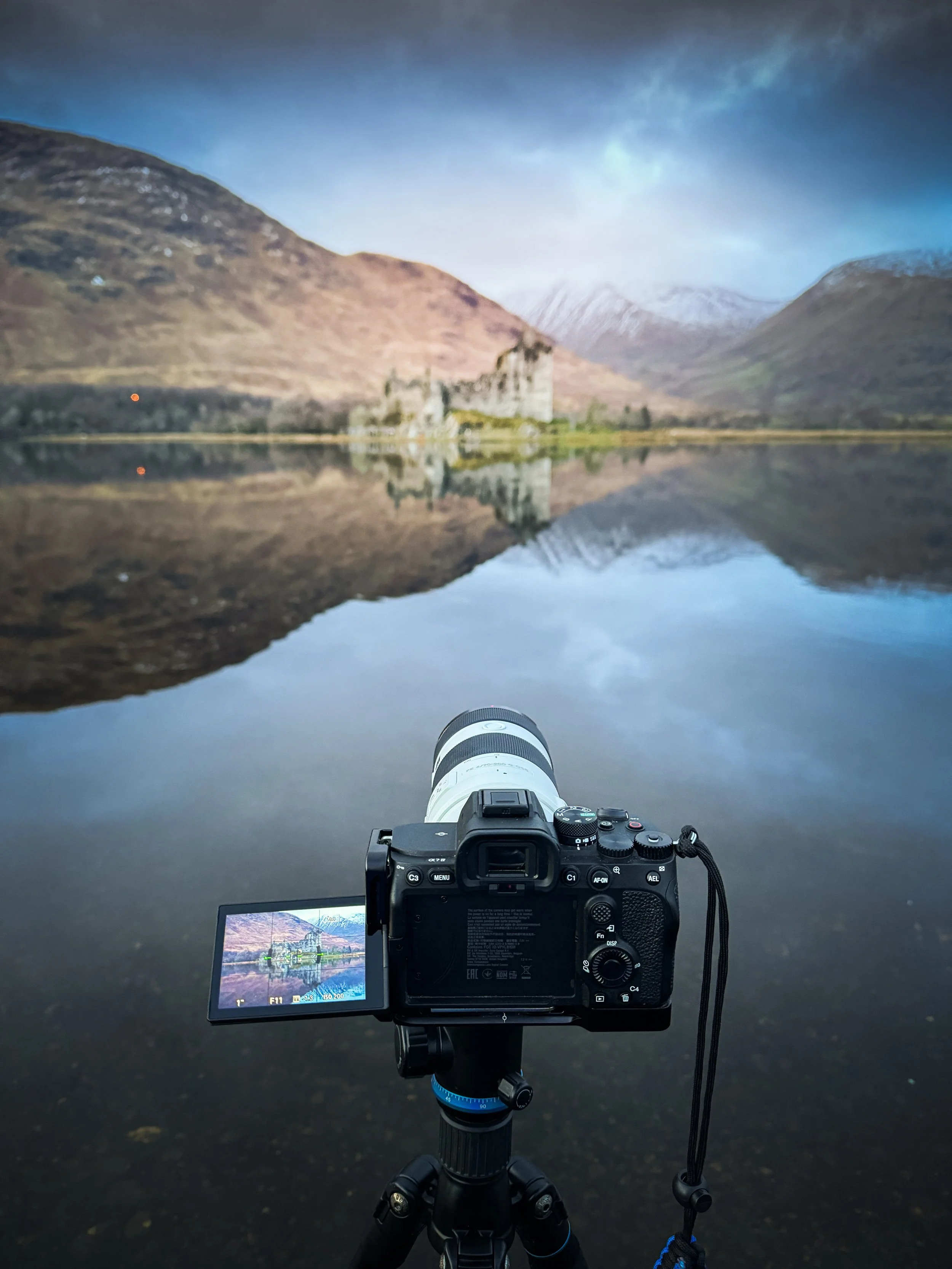 Camera on tripod capturing a landscape mirror reflected in a calm lake with mountains and a castle in the background.