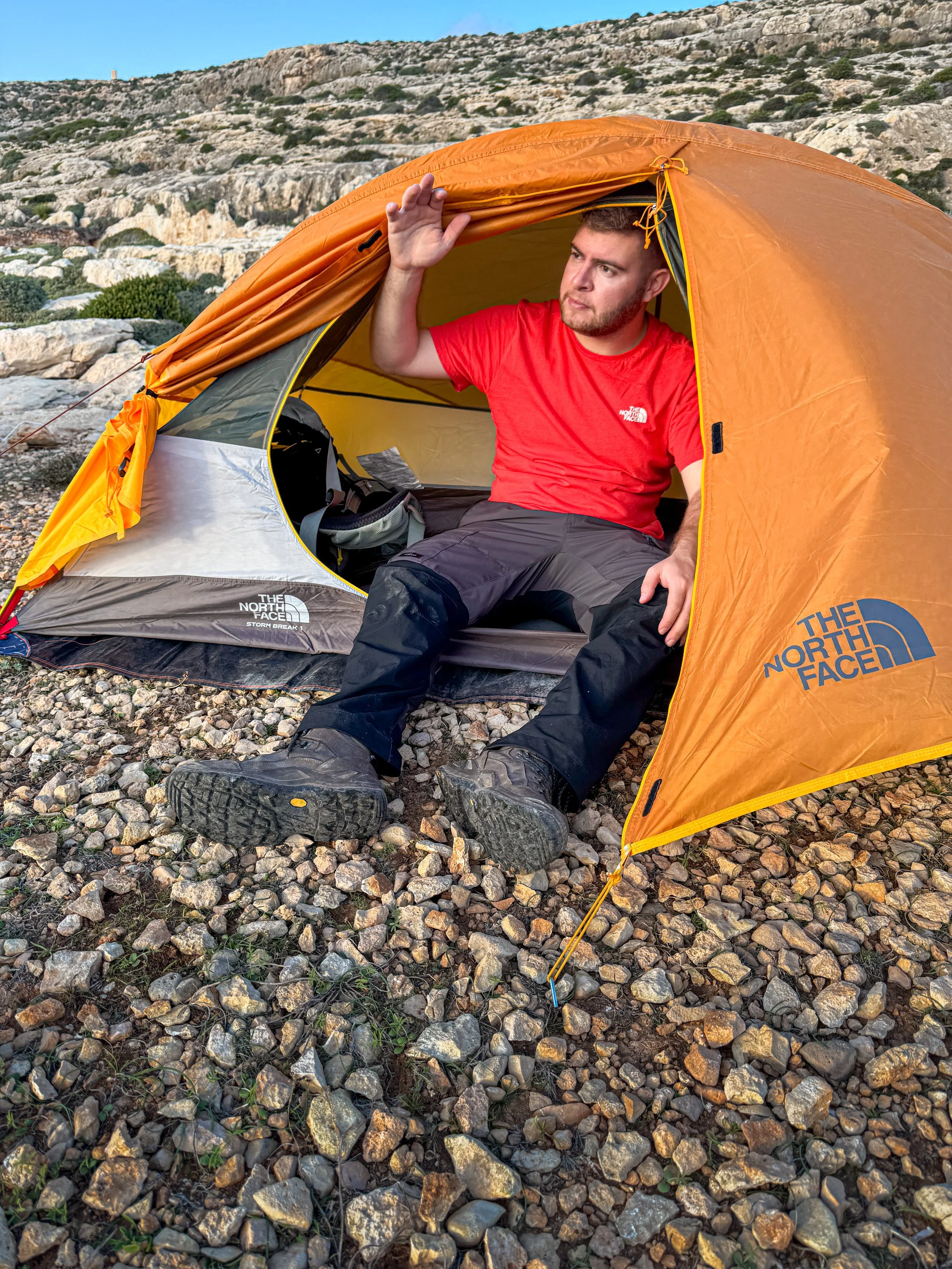 A man sitting inside a bright orange North Face tent on rocky terrain, wearing a red shirt and black pants, with a mountainous landscape in the background.