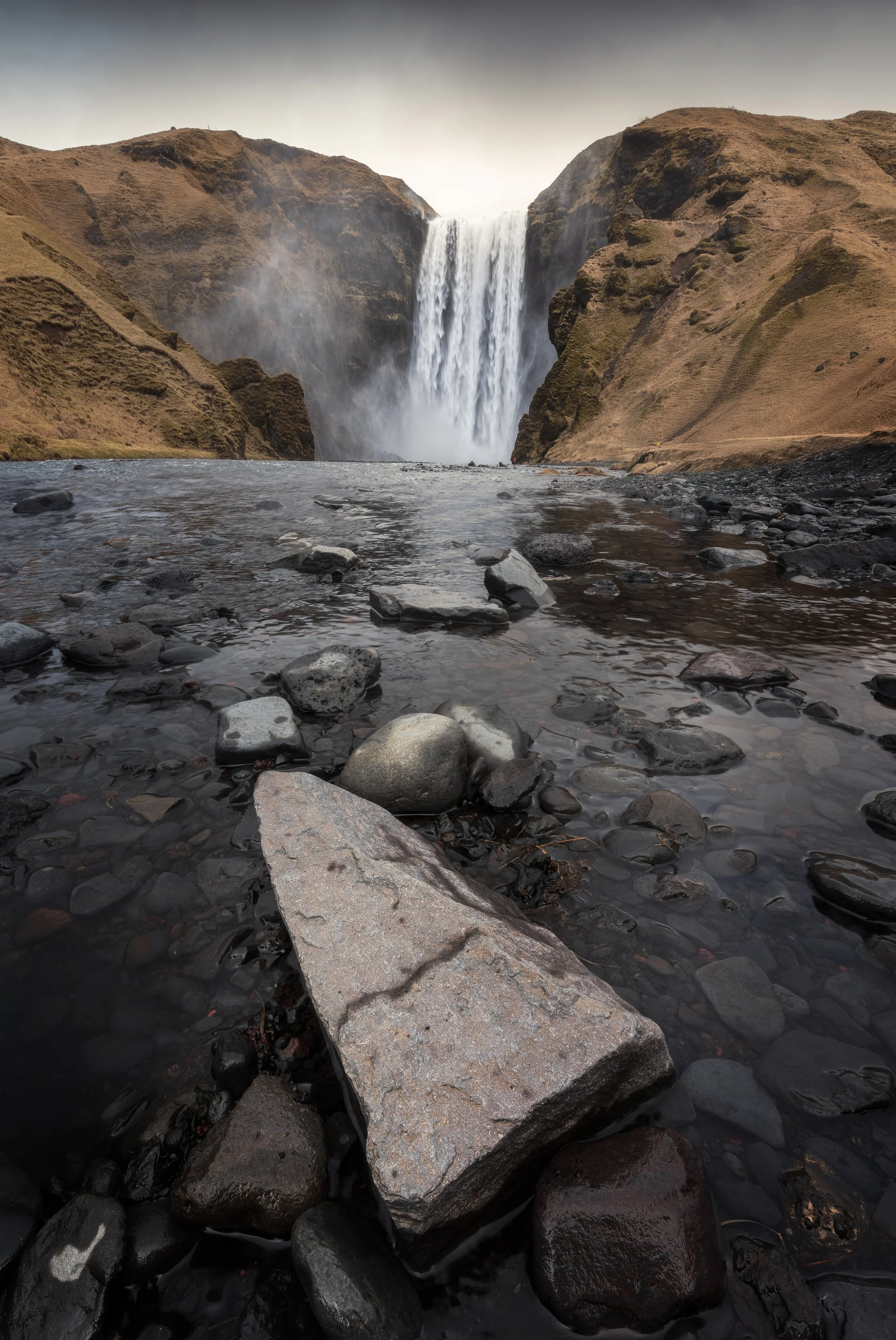 View of a waterfall with water cascading down cliffs into a shallow rocky river in a rugged landscape.