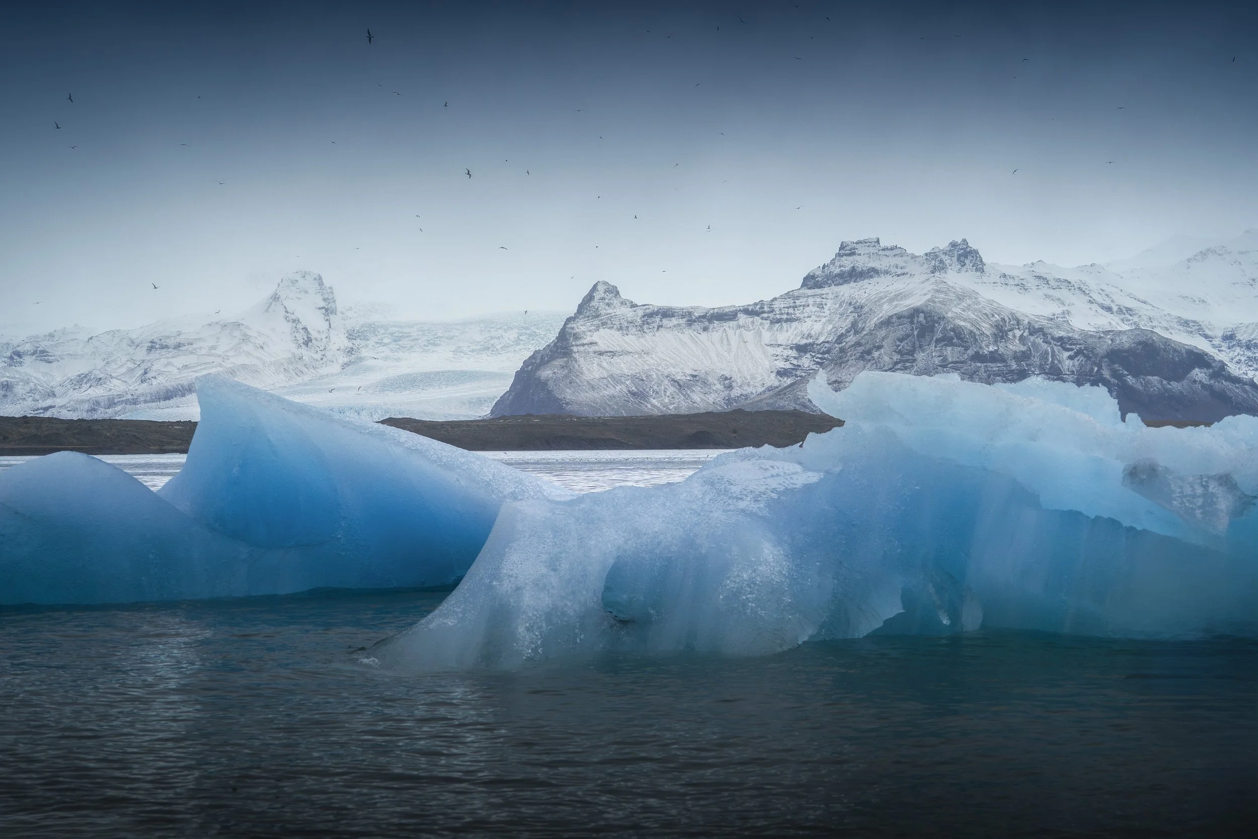 Icebergs floating in a body of water with snow-covered mountains and a cloudy sky in the background.