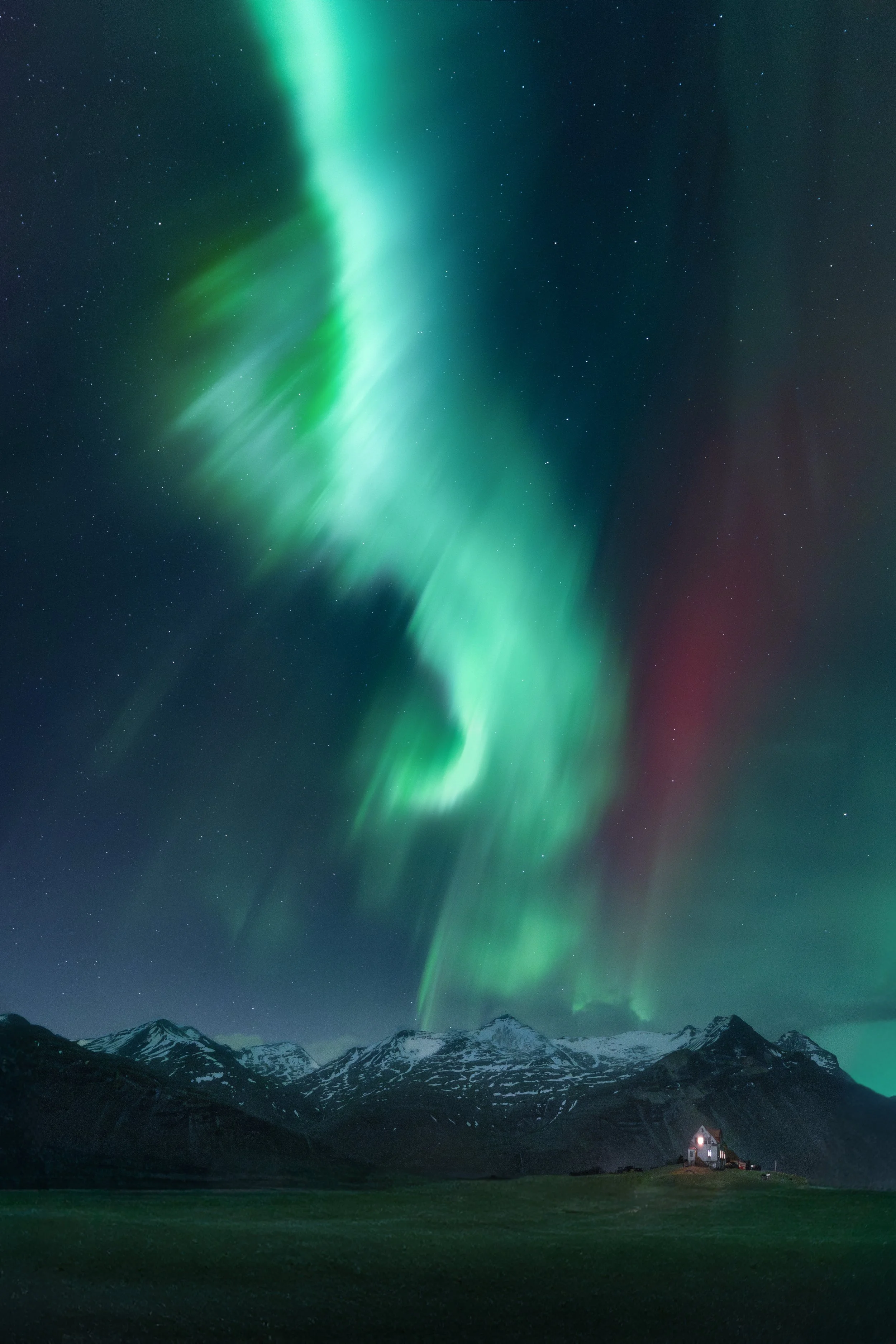 Northern lights over snow-capped mountains at night with a small, lit house in the foreground