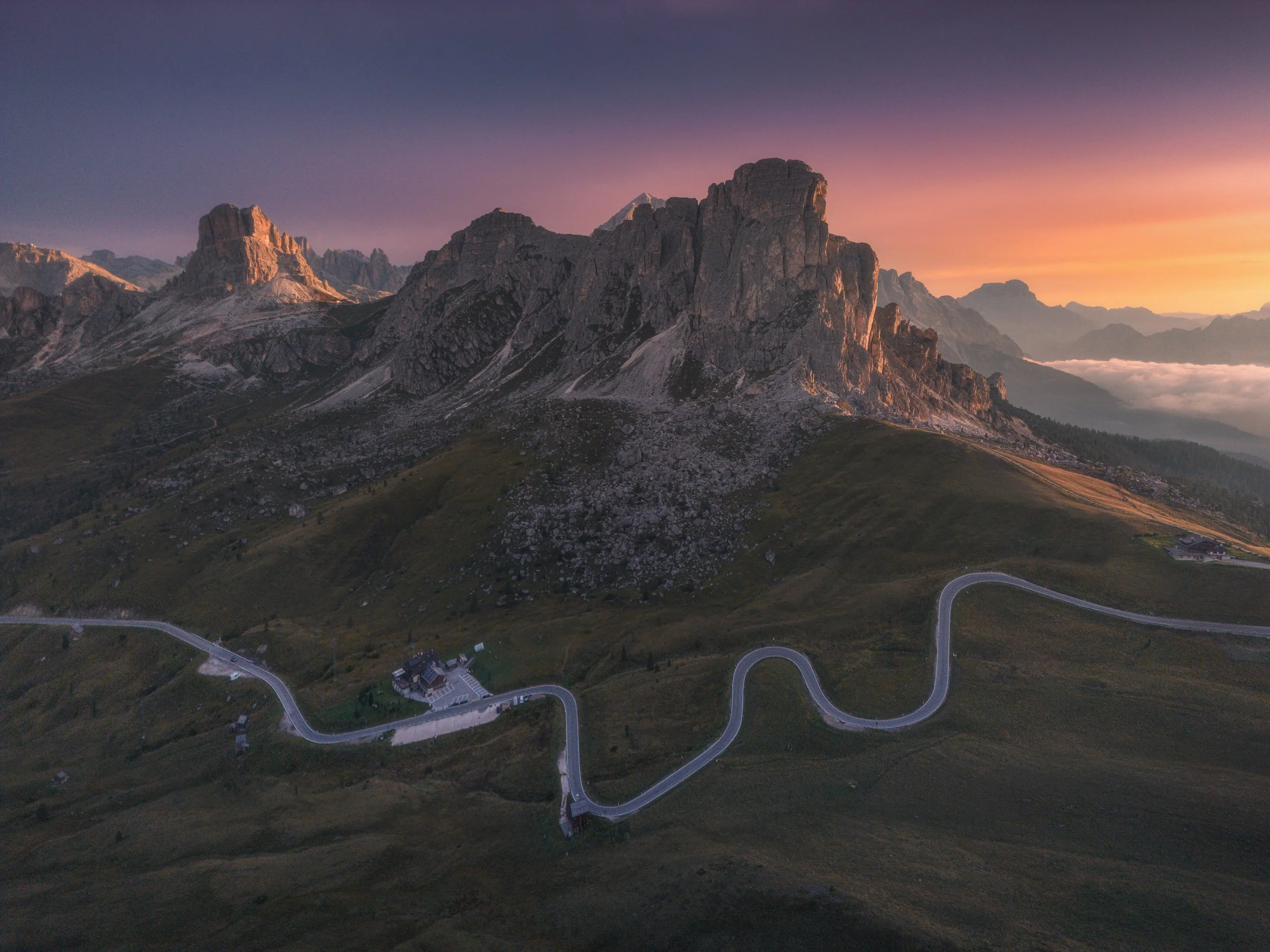 Mountain landscape at sunset with winding road and grassy slopes in the foreground.