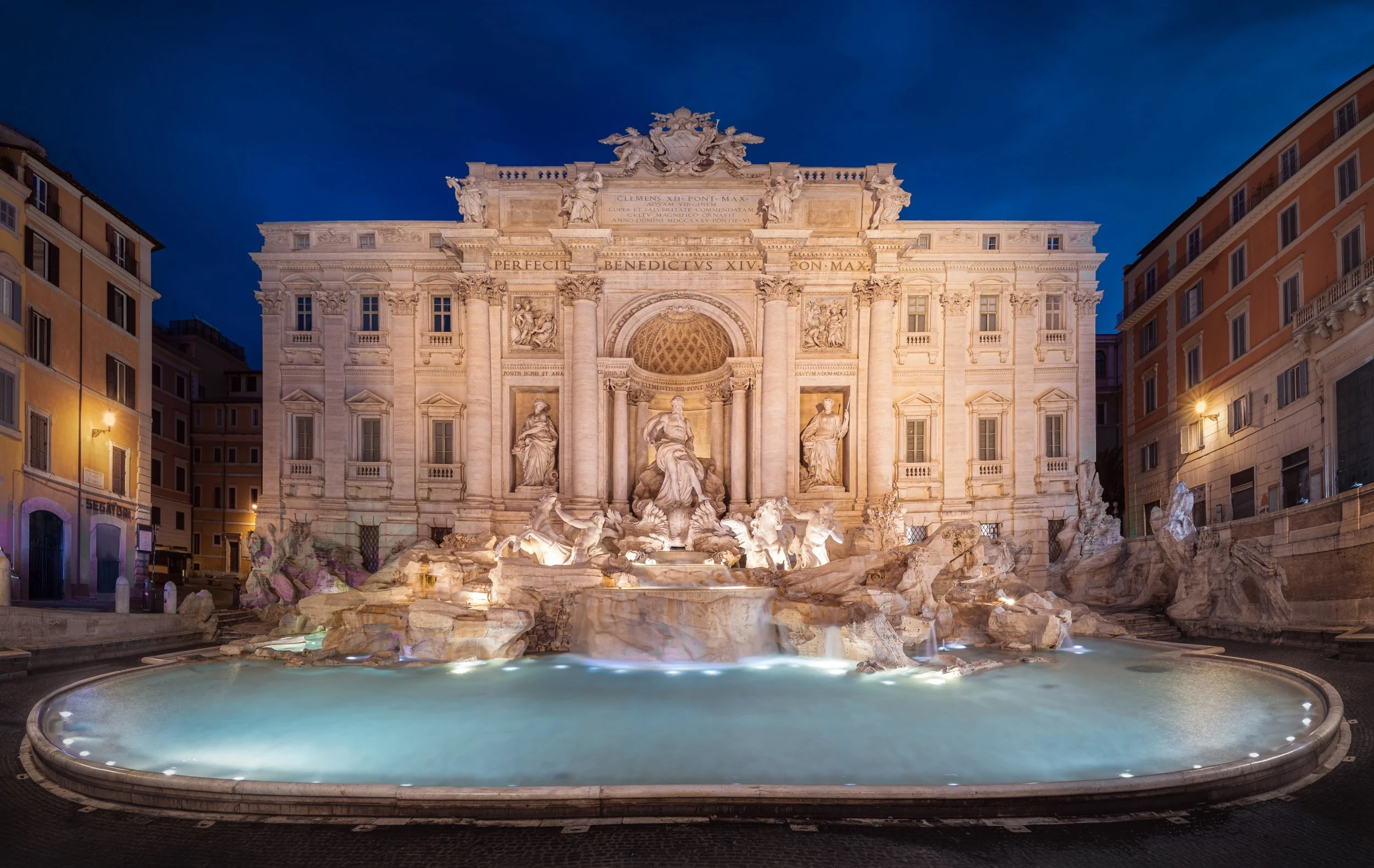 Night view of the ornate Trevi Fountain in Rome, Italy, illuminated and surrounded by buildings.