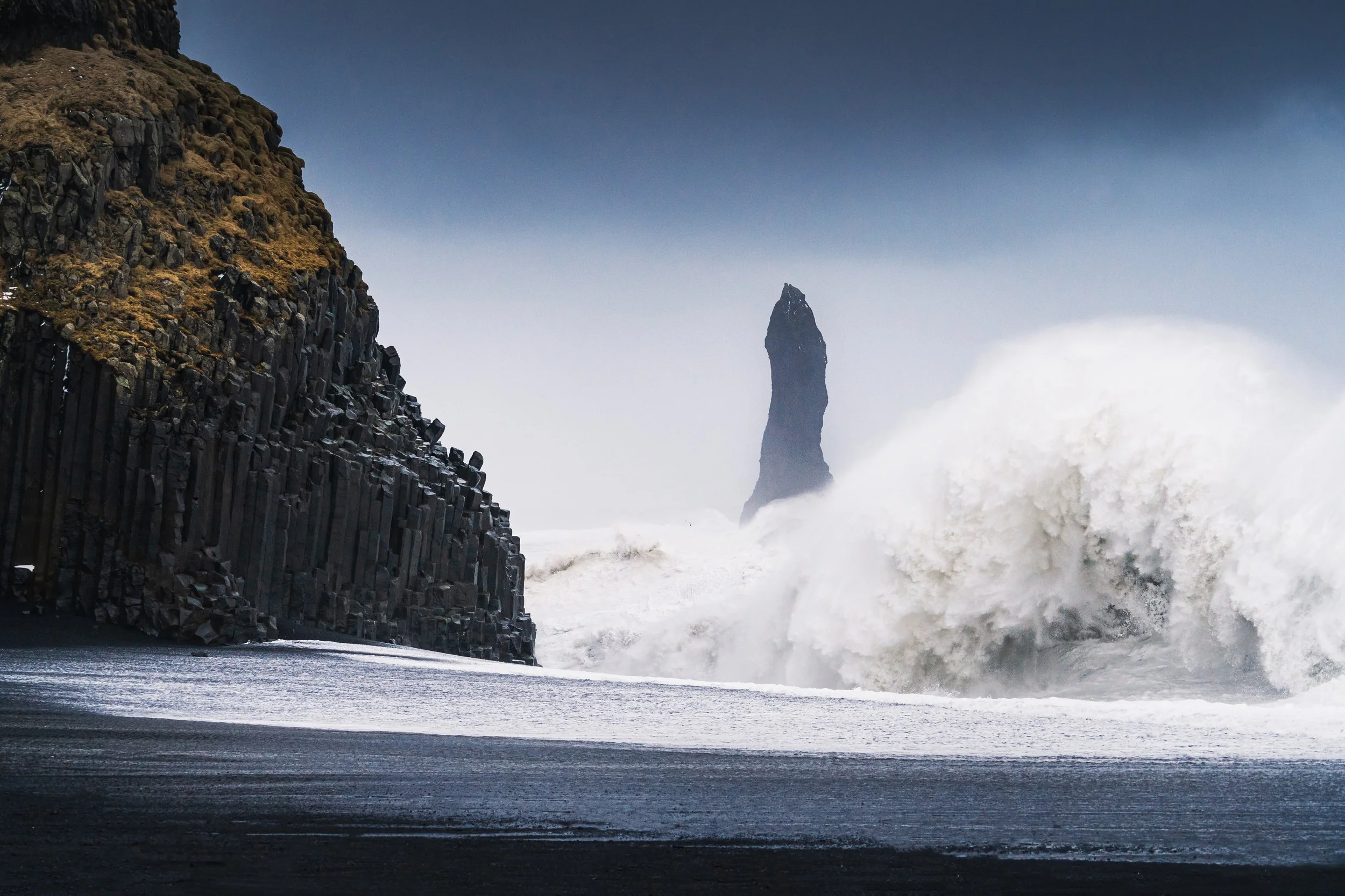 Ocean waves crashing against black volcanic rock cliffs with a tall sea stack in the distance under a cloudy sky.
