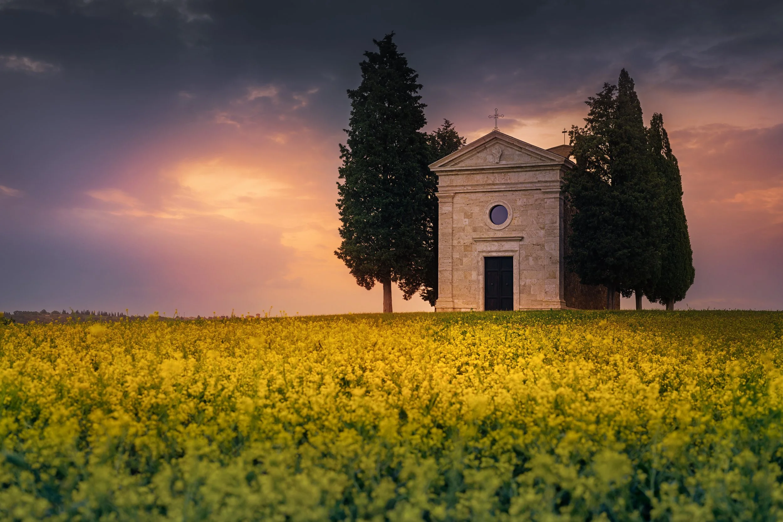 A small stone chapel surrounded by tall trees, set against a sunset sky, with a field of yellow flowers in the foreground.
