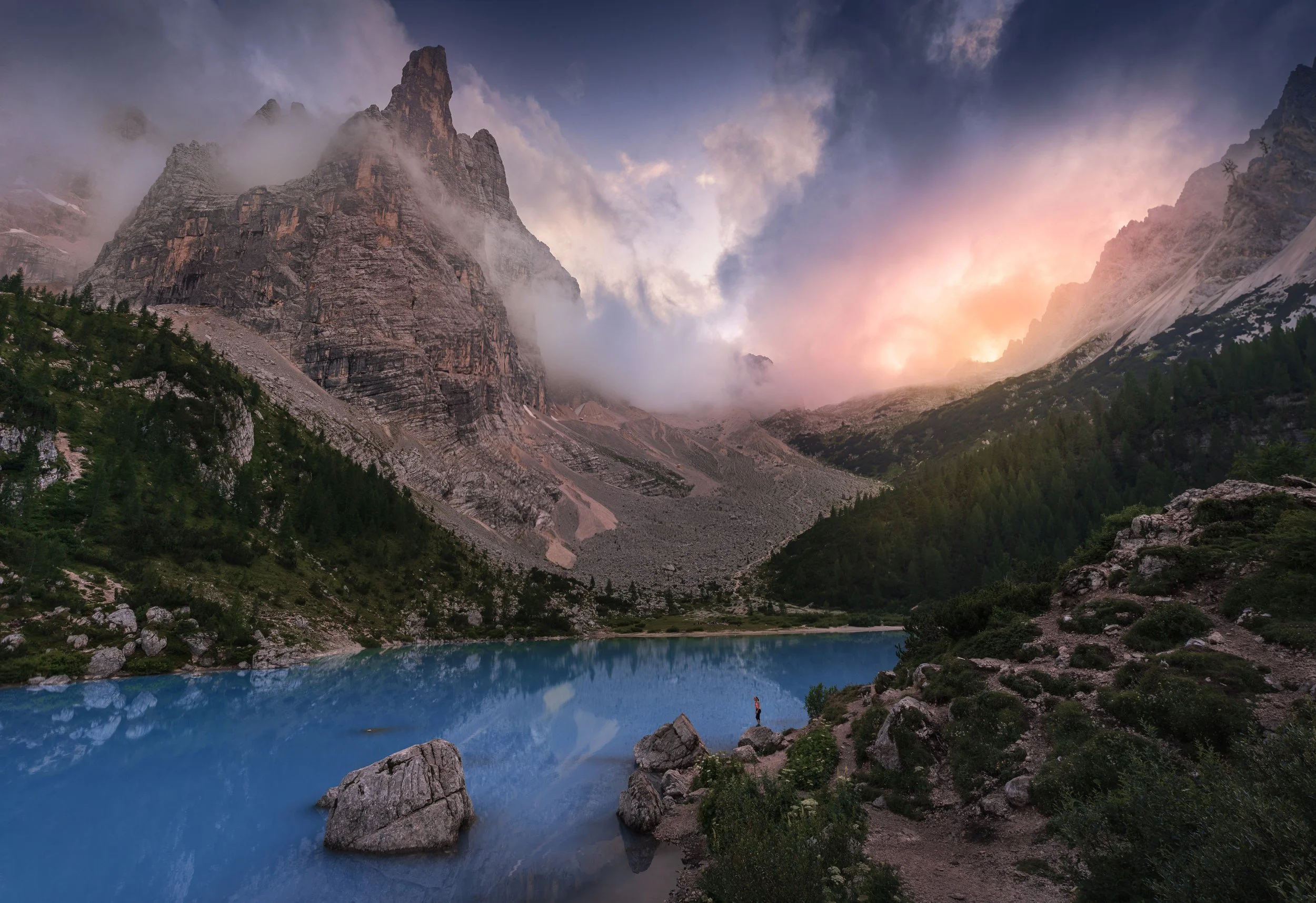 A mountain landscape at sunset with a large rocky peak, mist, a blue lake in the foreground, and a solitary hiker standing on rocks by the lake.