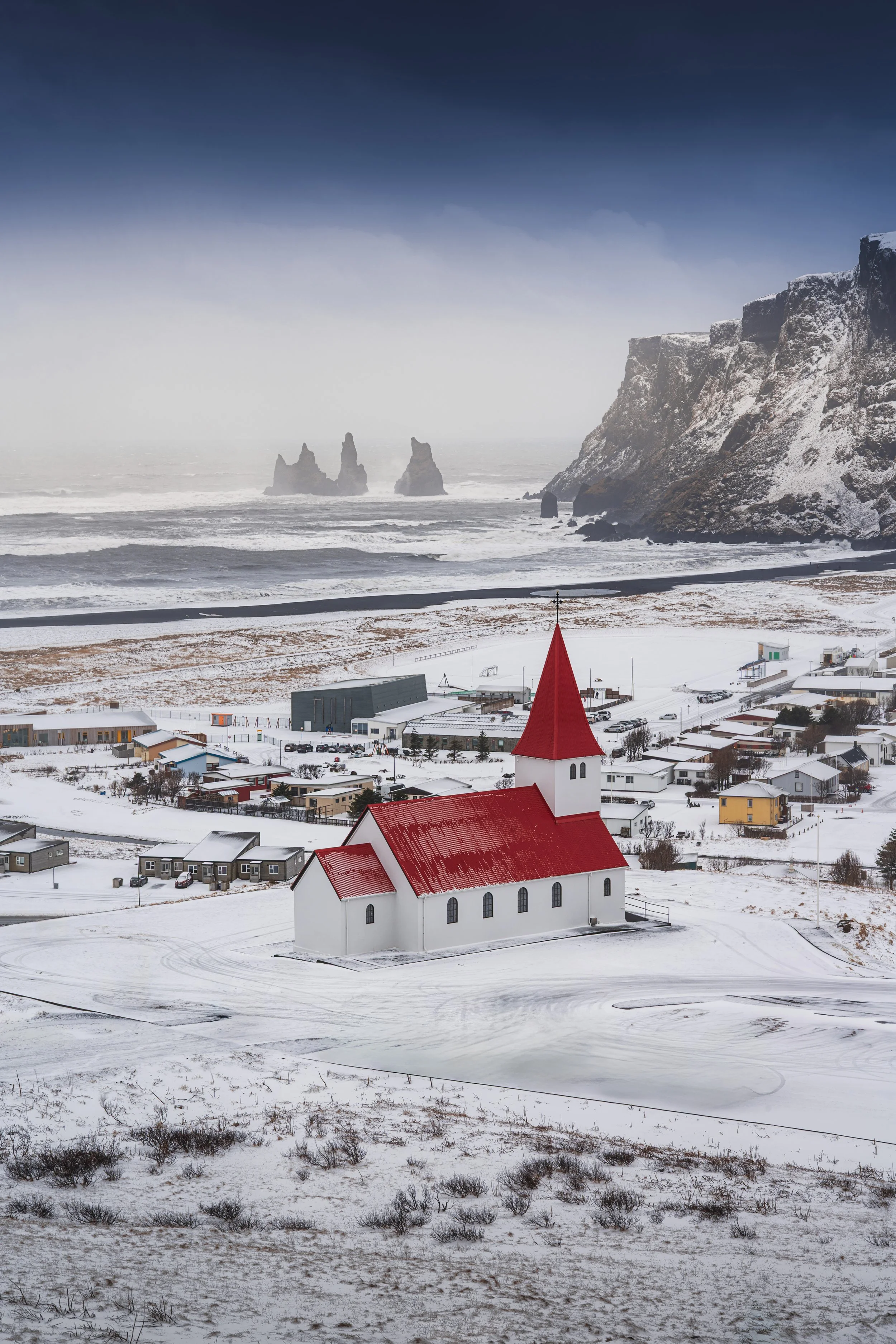 A snow-covered small white church with a red roof and steeple is situated on a hill overlooking a snowy town by the coast, with large rocky formations and cliffs in the ocean in the background.