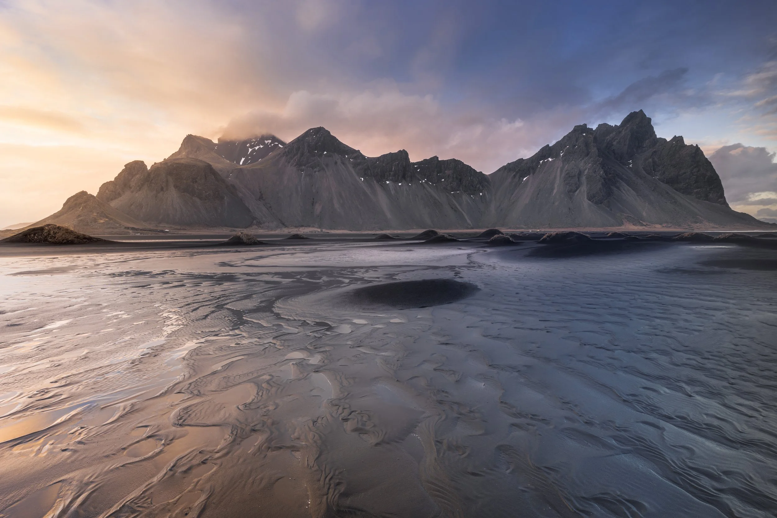 Mountain landscape with dark, rugged peaks, cloudy sky, and textured sandy shore in the foreground.