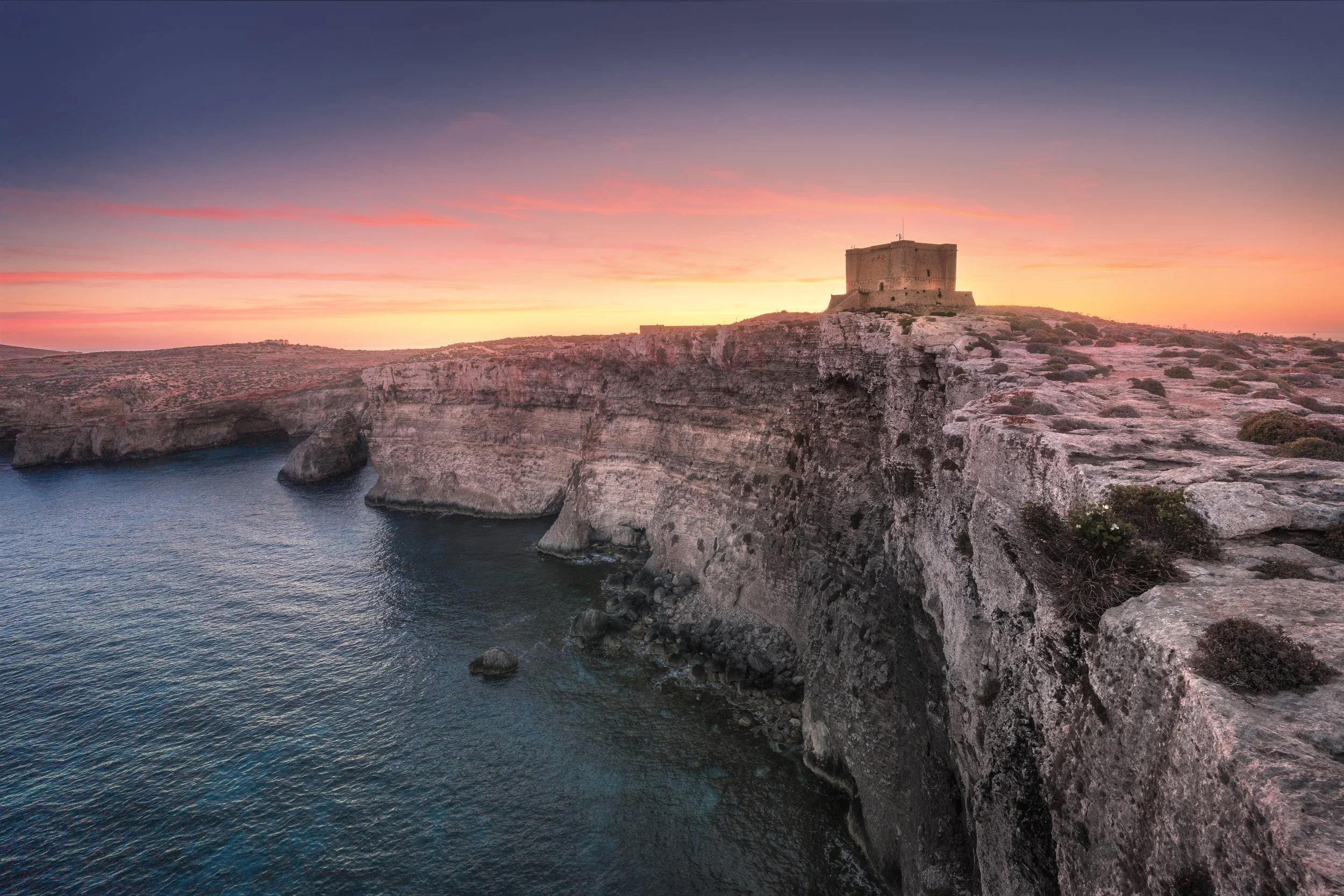 Sunset over a rocky coastline with a historic fortress on a cliff.