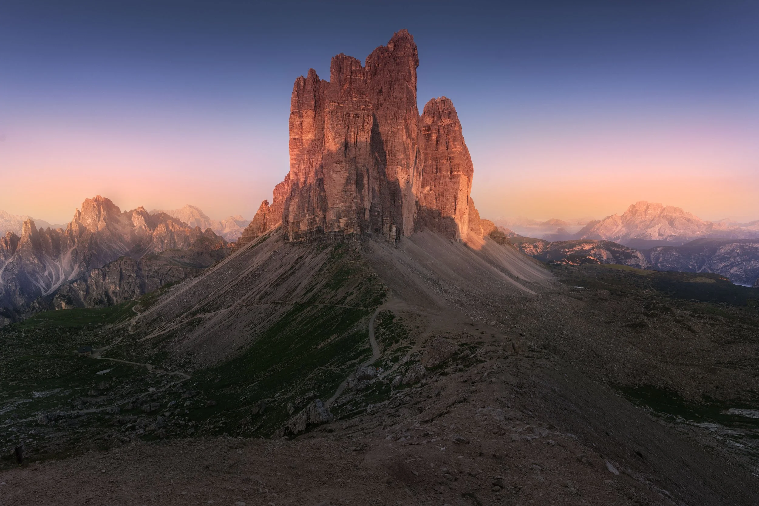 A tall, rugged mountain during sunset with a clear sky, surrounded by smaller mountains and green and rocky terrain.