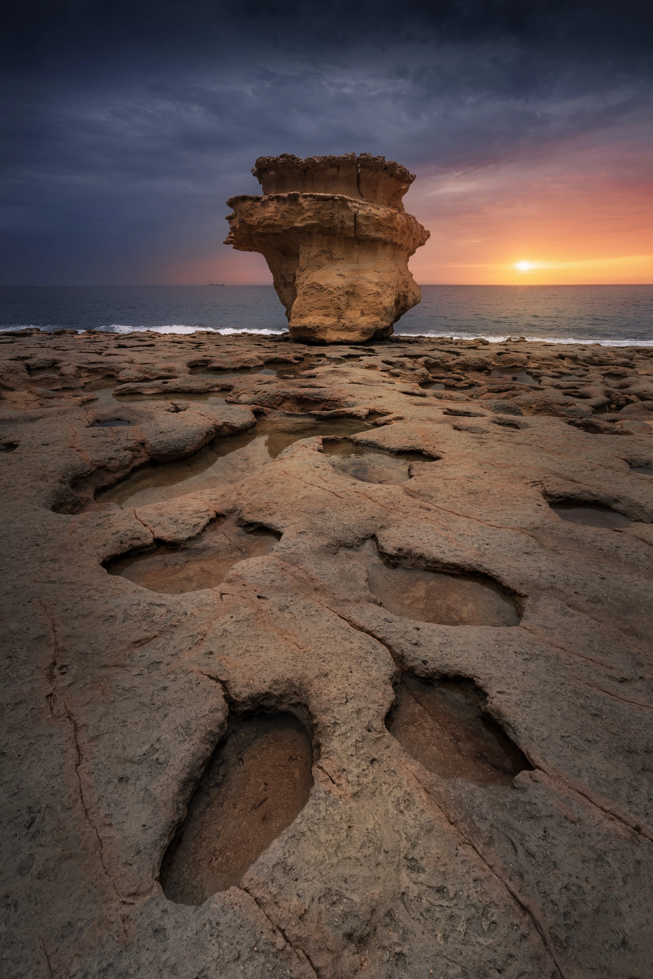 Sunset over the ocean with a large weathered rock formation on a rocky shoreline and tide pools in the foreground.