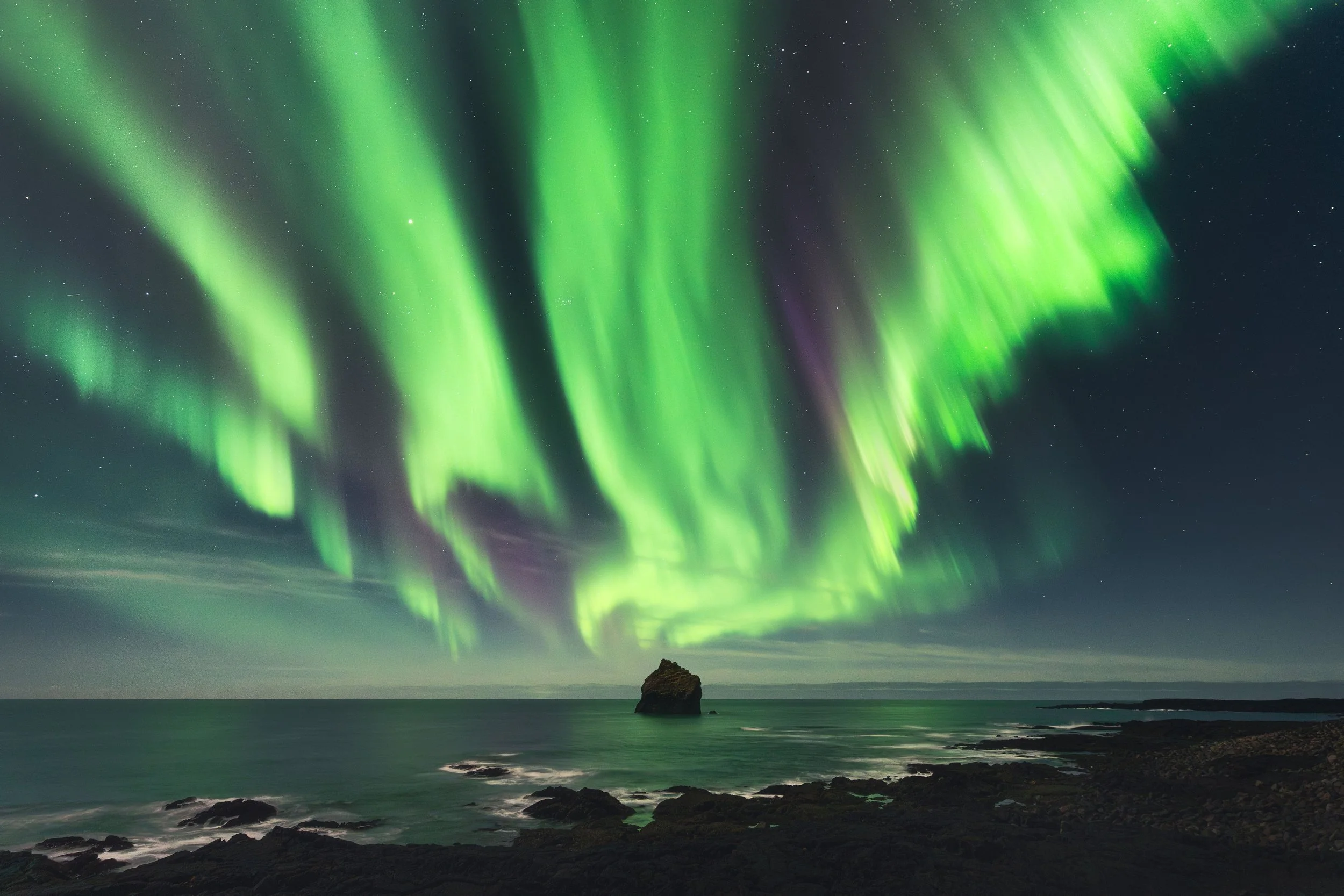 Northern Lights (aurora borealis) over a rocky shoreline with a large rock in the water, clear night sky with stars.