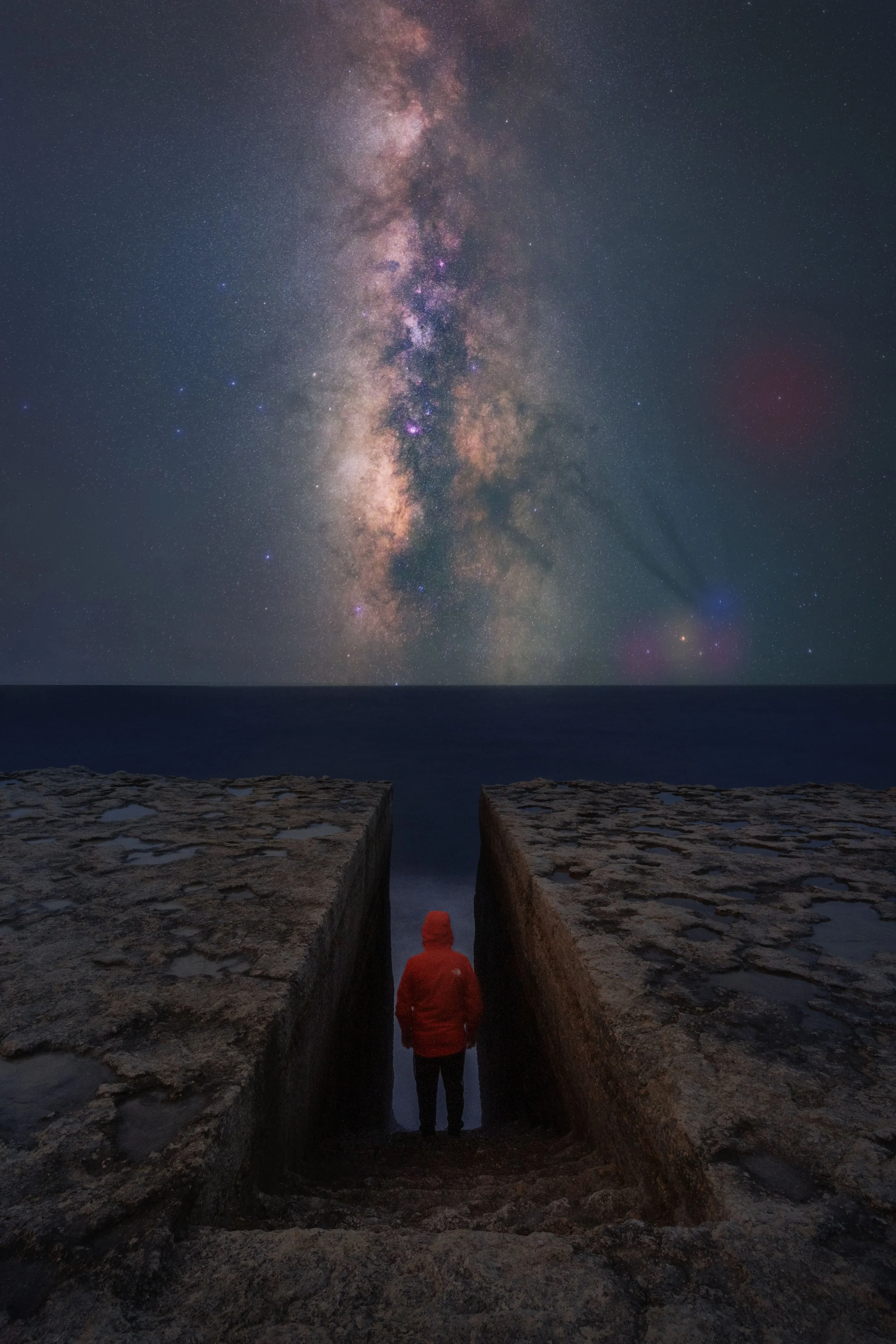 A person in a red jacket stands between two large stone walls, looking at the night sky filled with stars and the Milky Way galaxy over the ocean.