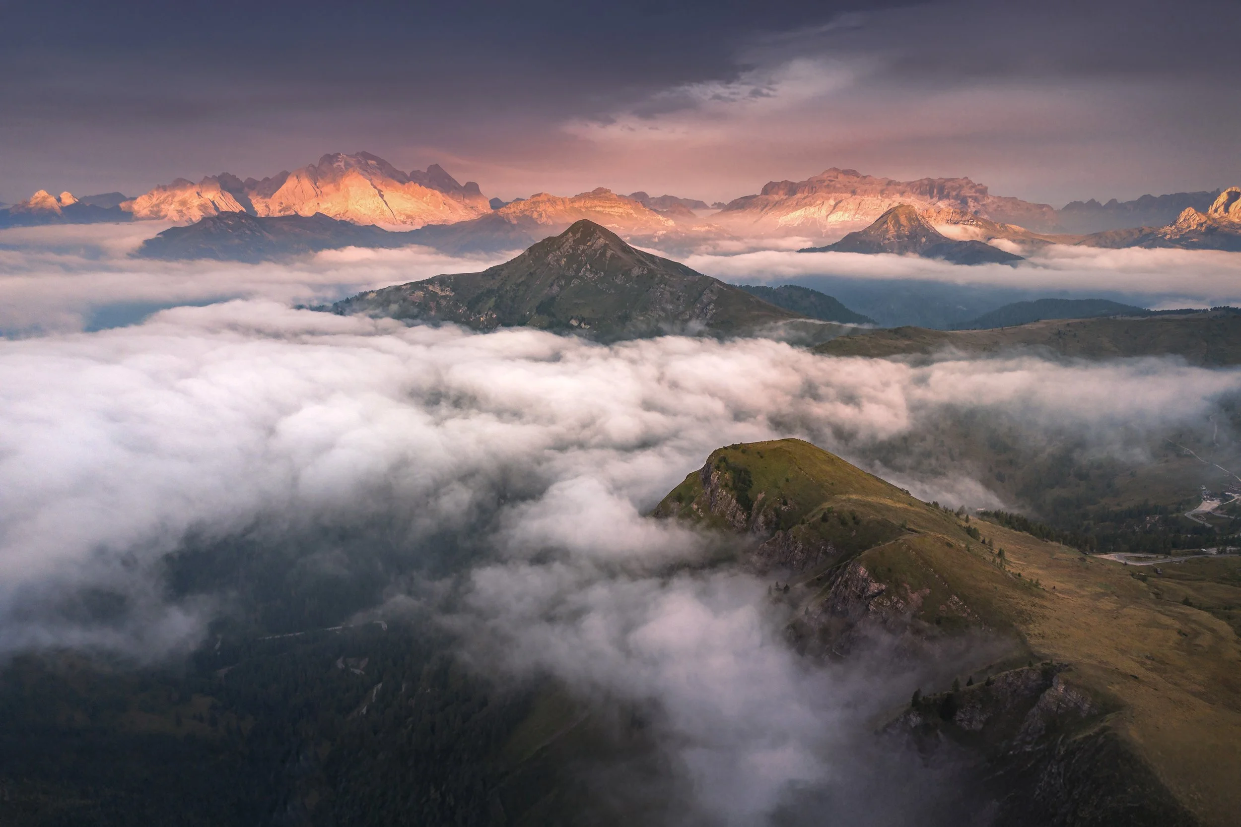 A mountain landscape at sunrise or sunset with peaks in the distance, clouds covering the lower elevations, and a rugged hillside in the foreground.