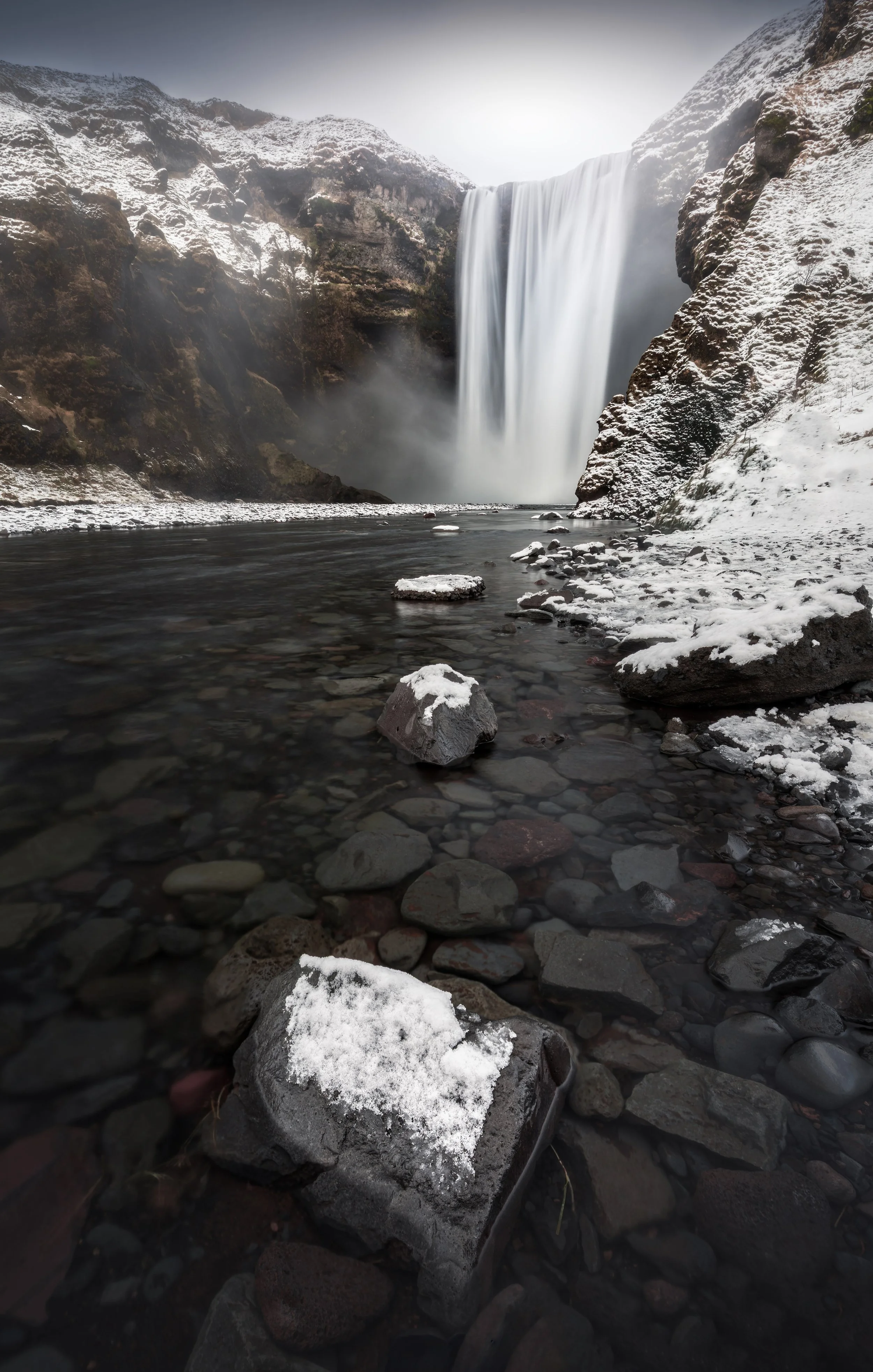 Snow-covered rocks and pebbles along a river in front of a large waterfall in a mountainous, snowy landscape.