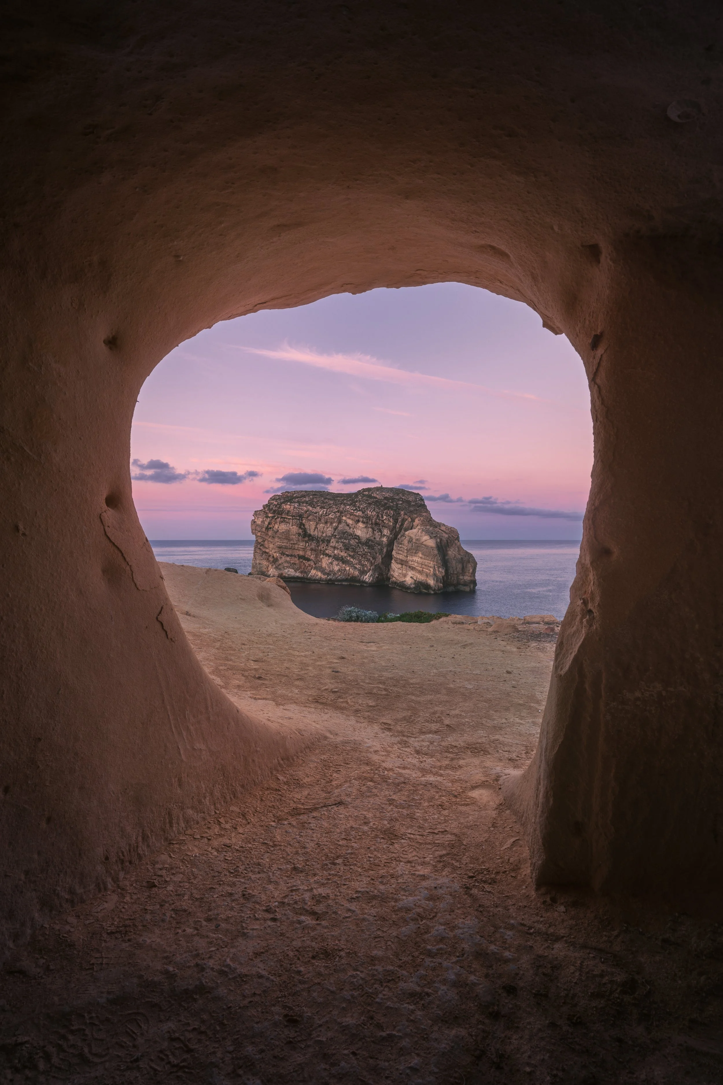 View of a large rocky island in the ocean, seen through a naturally formed rock window at sunset, with pink and purple sky and scattered clouds.