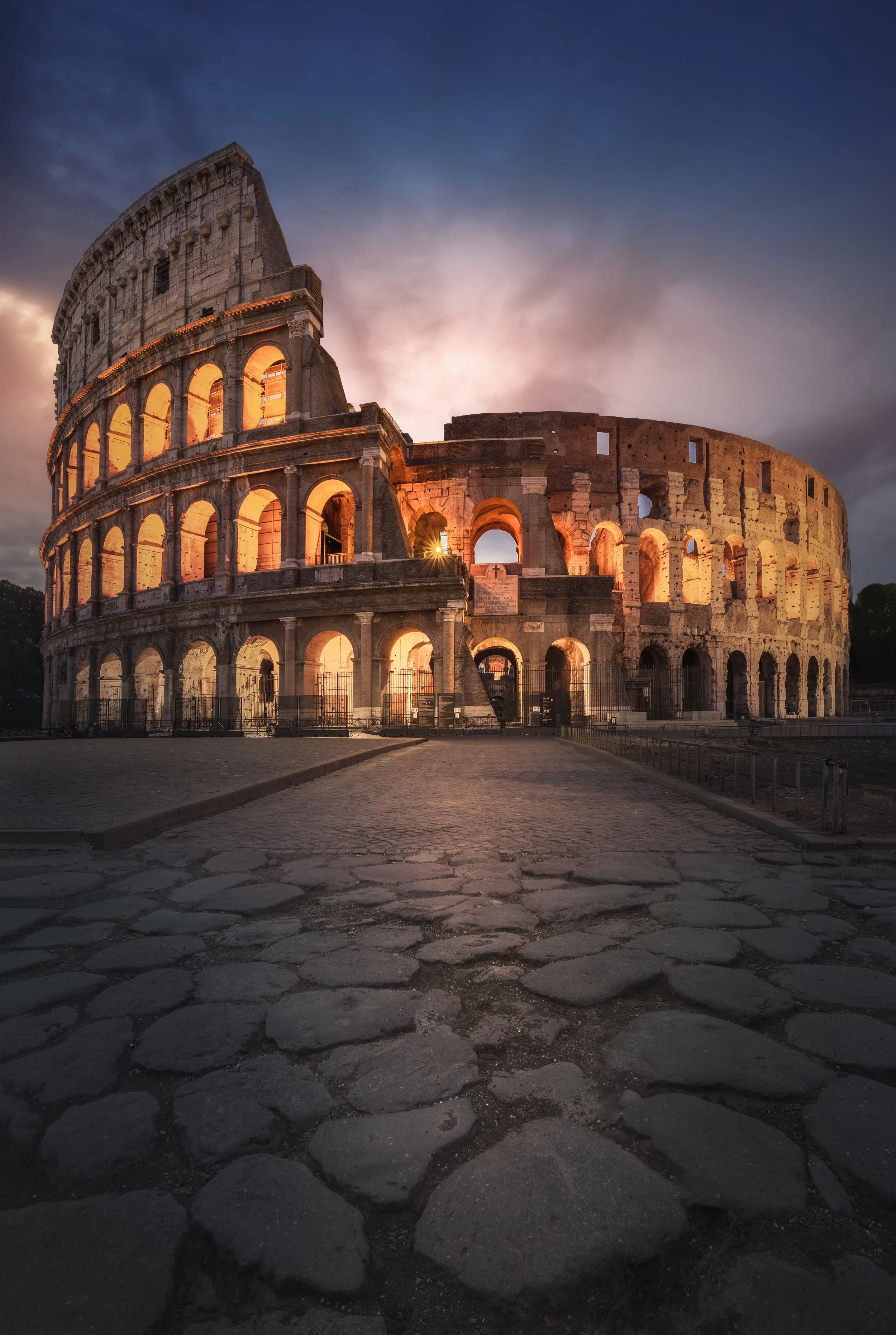The illuminated exterior of the Roman Colosseum at dusk with a cloudy sky in the background.