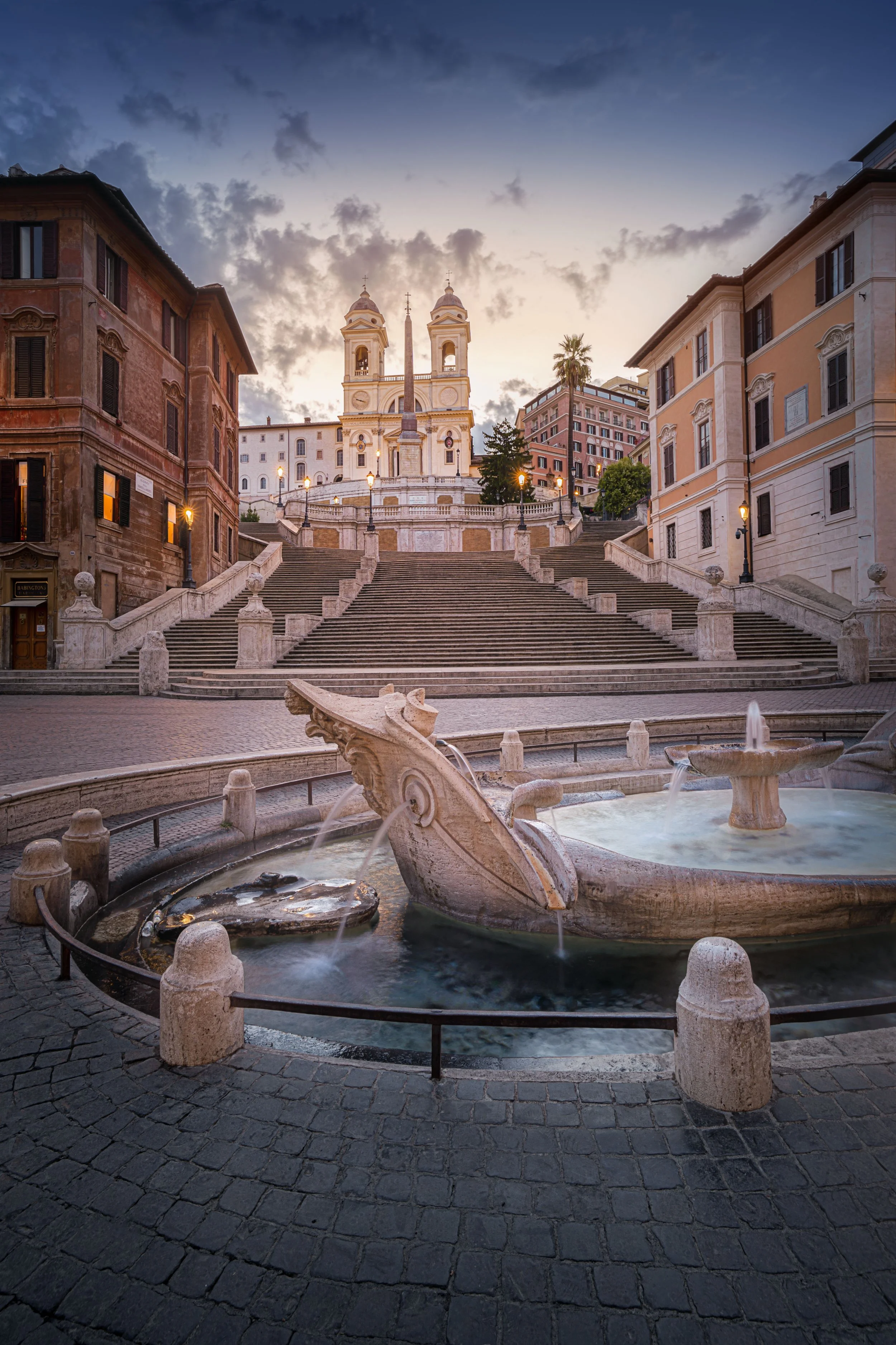 View of the Spanish Steps leading up to the Trinità dei Monti church in Rome, with a fountain featuring a dragon sculpture in the foreground, surrounded by historic buildings and a cloudy sky at sunset.
