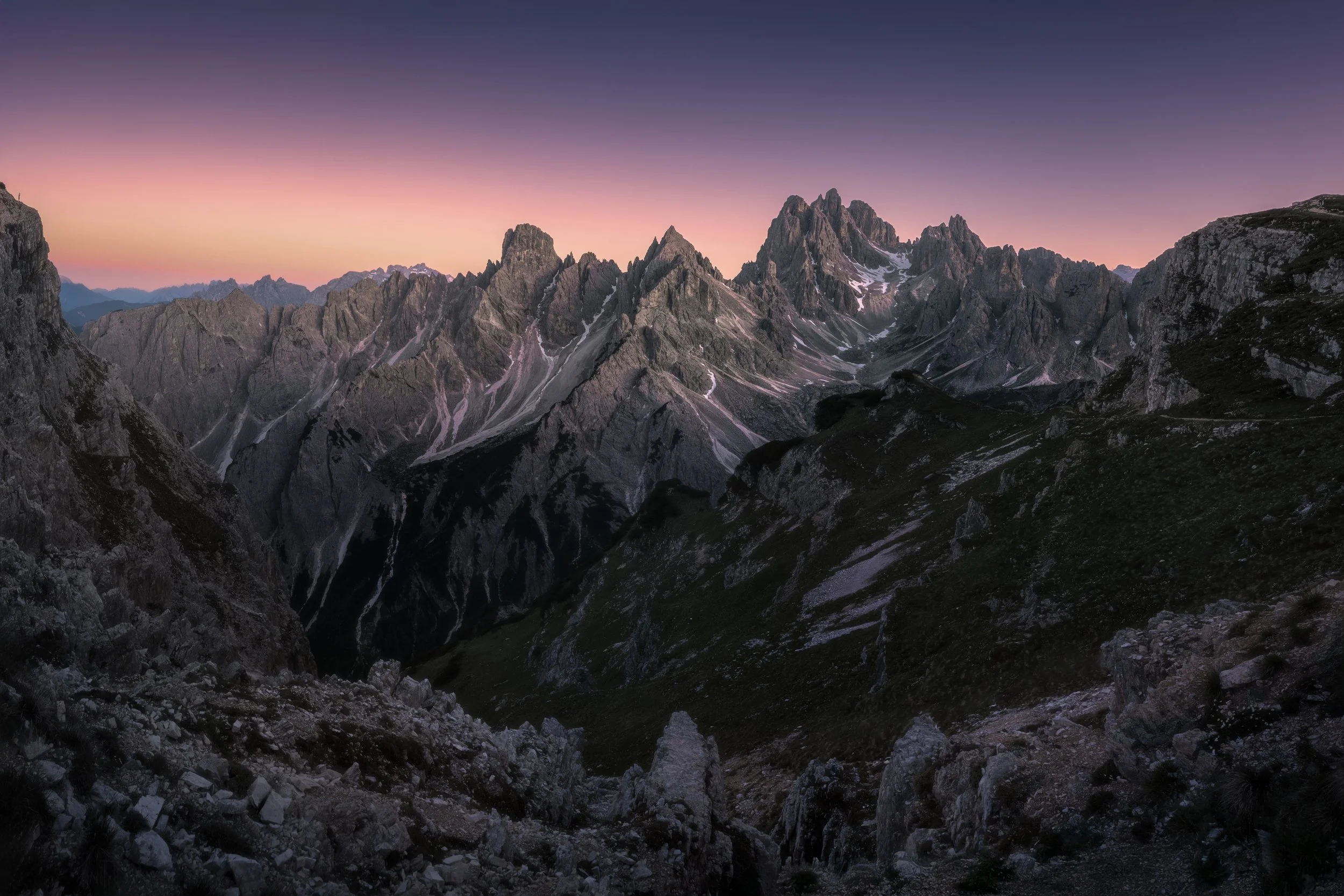 Mountain landscape at dawn with rocky peaks, some snow patches, and a colorful sky transitioning from pink to purple.