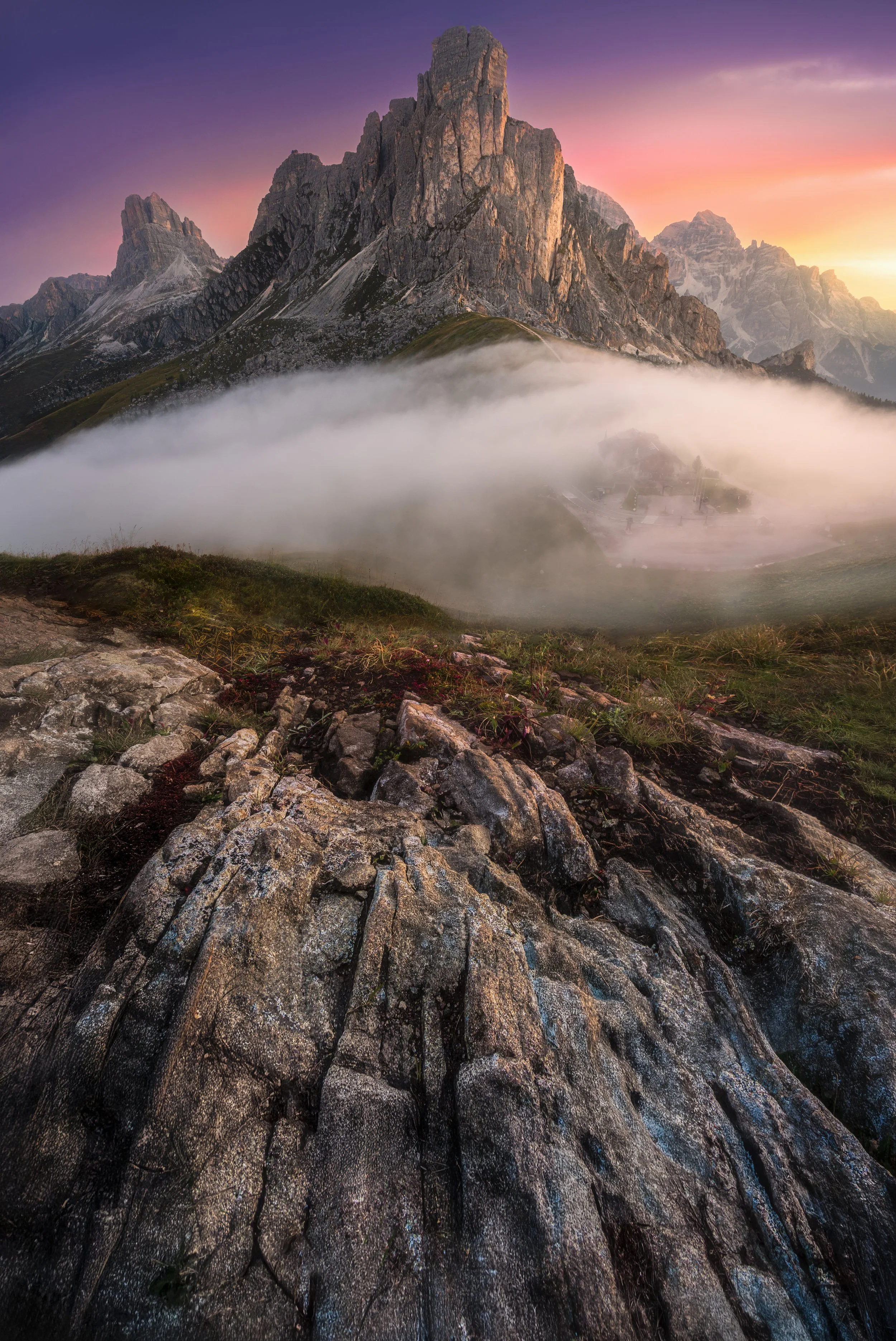 A mountain landscape at sunrise with a rocky foreground, mist in the mid-ground, and tall rugged peaks in the background, under a colorful sky.