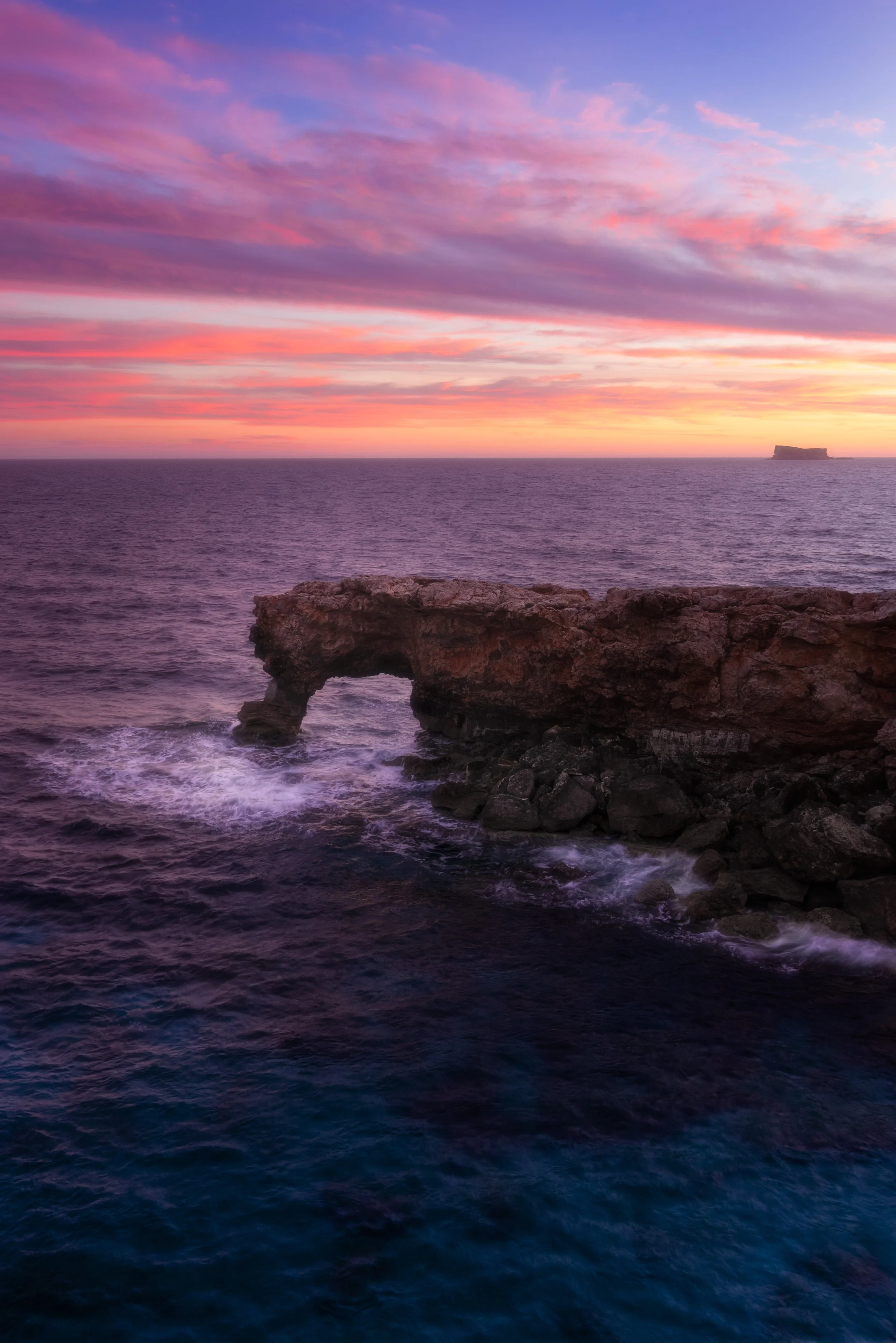Sunset over the ocean with pink and purple clouds, a natural rock arch at the shore, and a distant small island on the horizon.