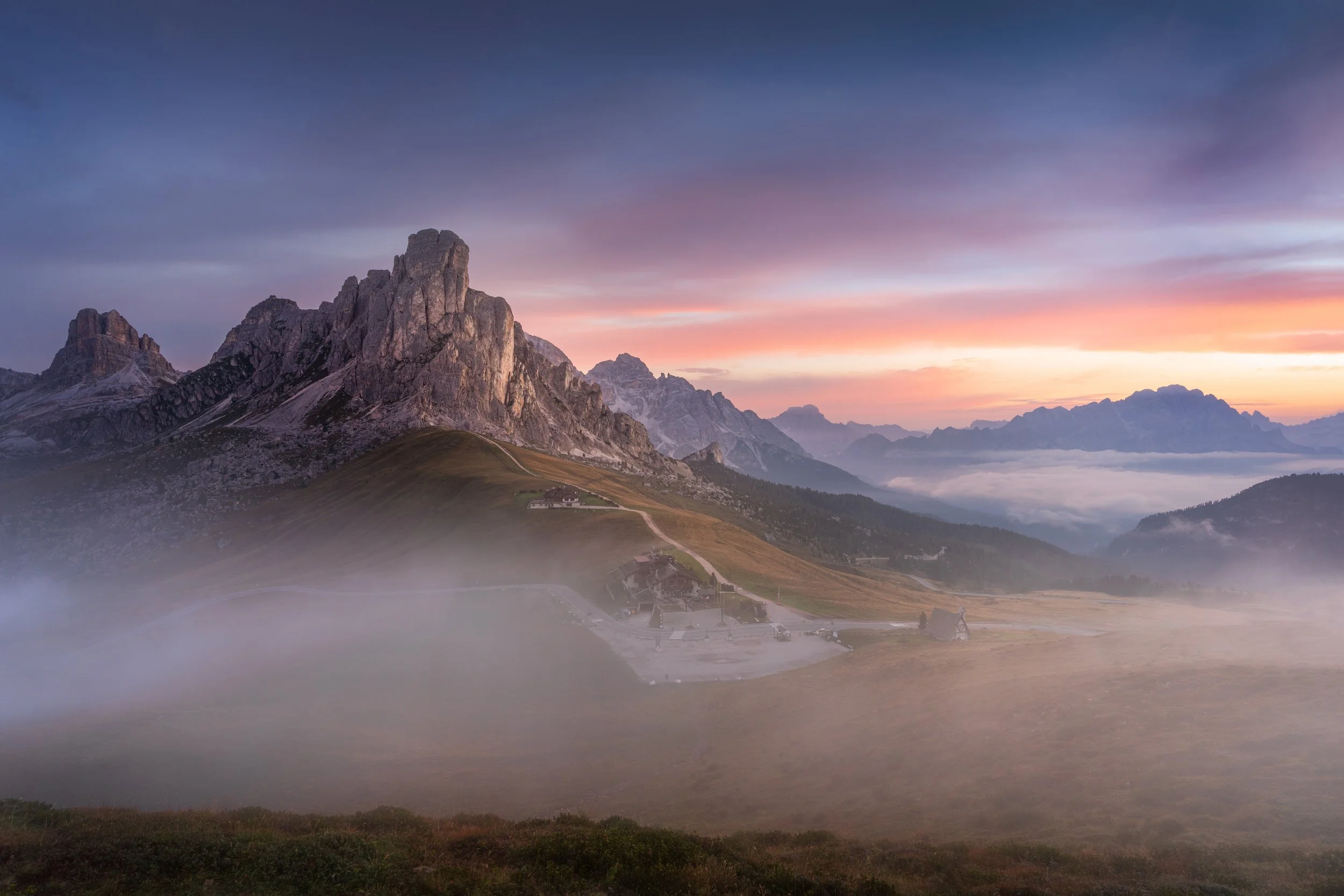 Mountain landscape during sunrise with fog in the valleys, rugged peaks, and a small village with buildings and winding roads.