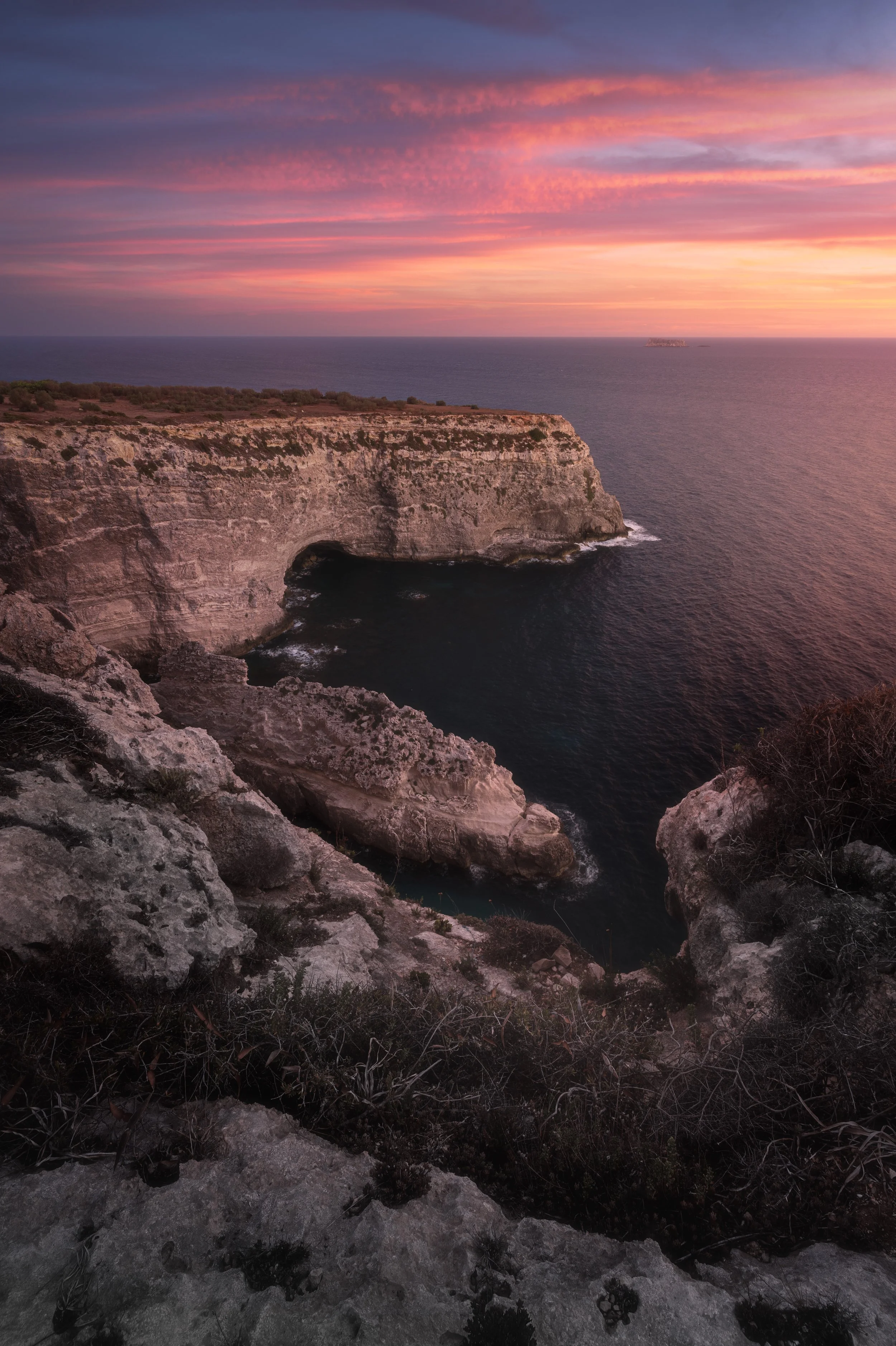Sunset over rocky coastal cliffs with the ocean below and a pink, purple, and orange sky.