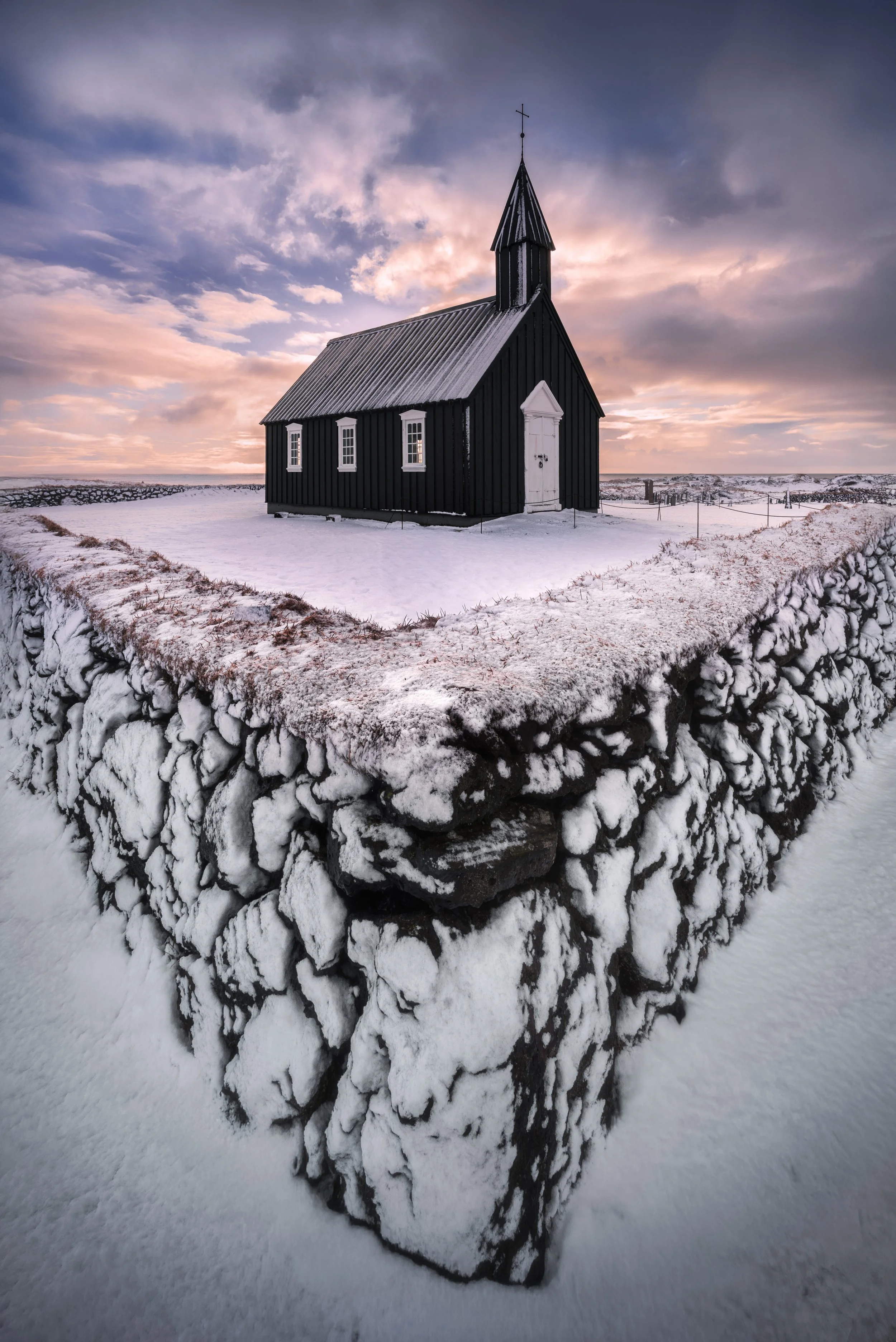 A small black wooden church with a white door and windows, surrounded by snow, in a rural landscape at sunset with a cloudy sky.