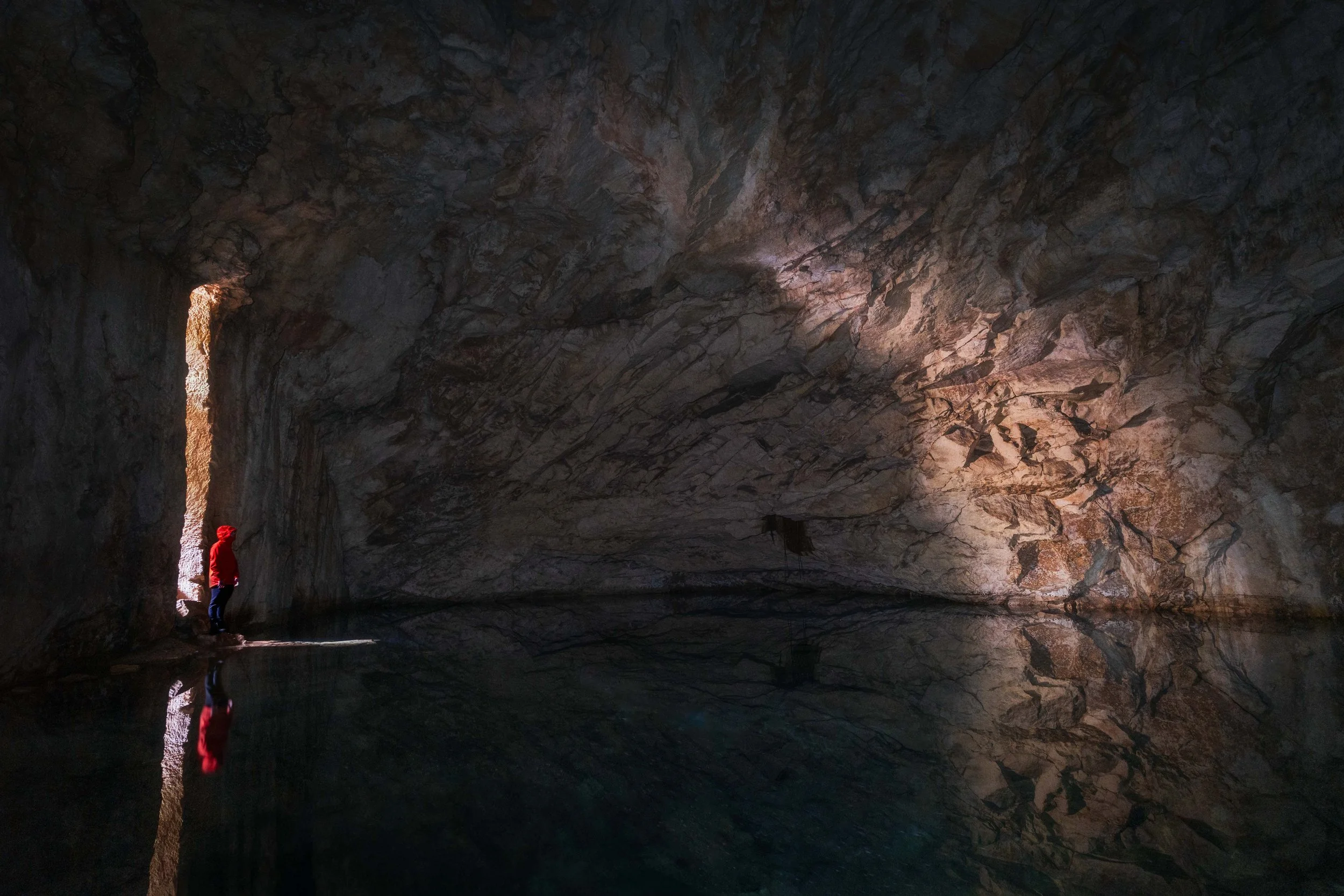 A person in a red jacket standing at the edge of a cave with water, looking into the dark interior and reflected in the water.